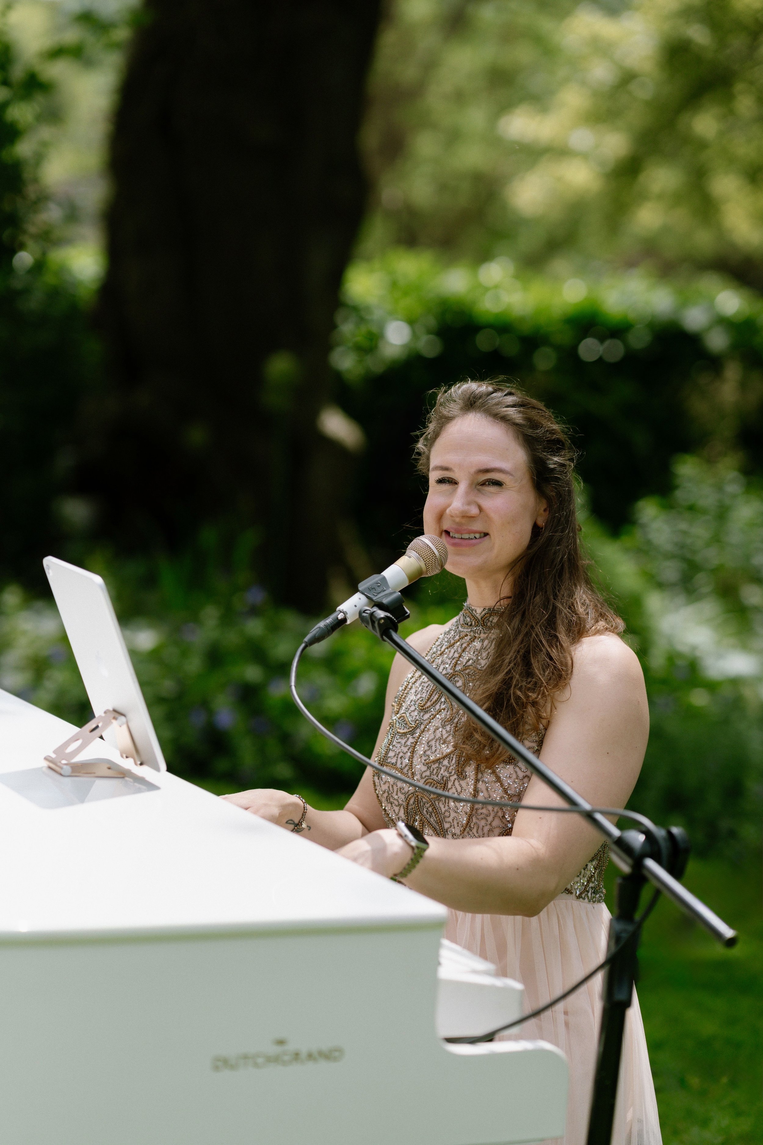 Kim Brooklyn singing and playing a white piano at an outdoor wedding