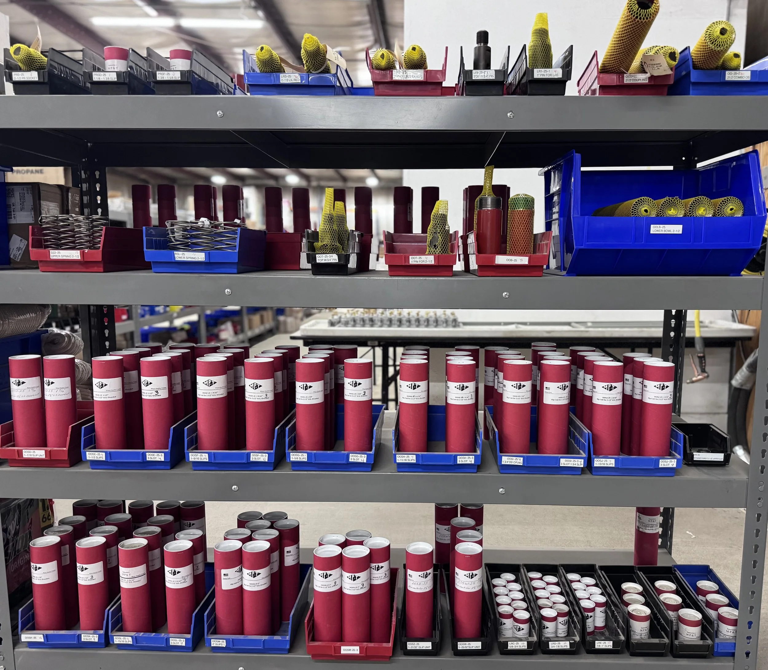Shelves filled with red and black cans and small containers, many with white labels, and yellow mesh tape rolls on the top shelf in a hardware store.