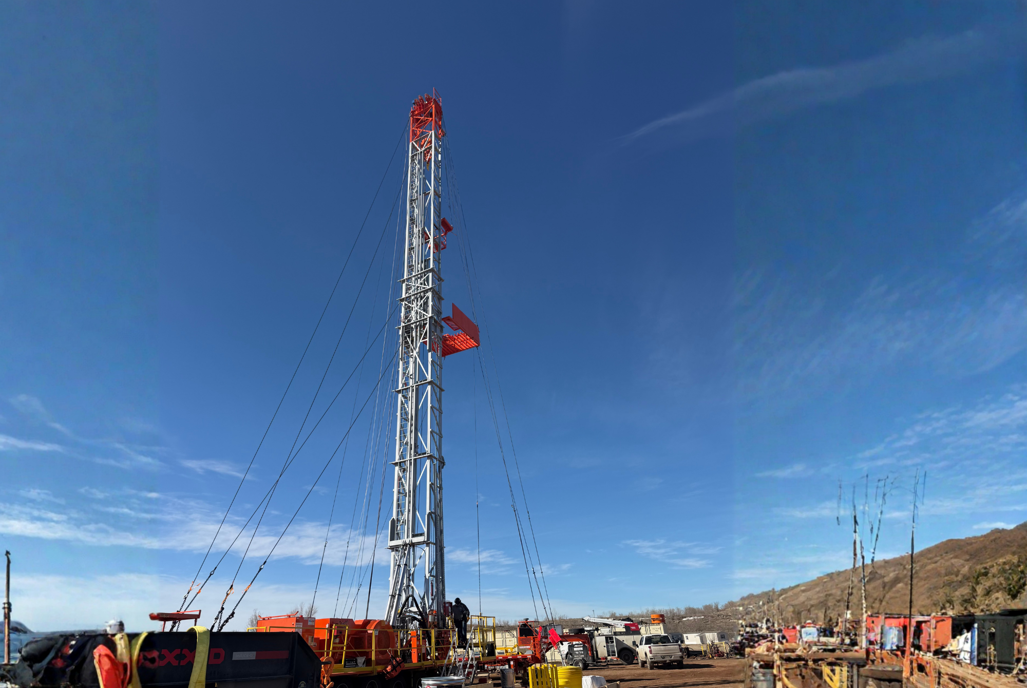 A tall oil drilling rig tower with red and white sections stands against a blue sky. Several vehicles, equipment, and people are at the base of the rig in a rural area with mountains in the background.