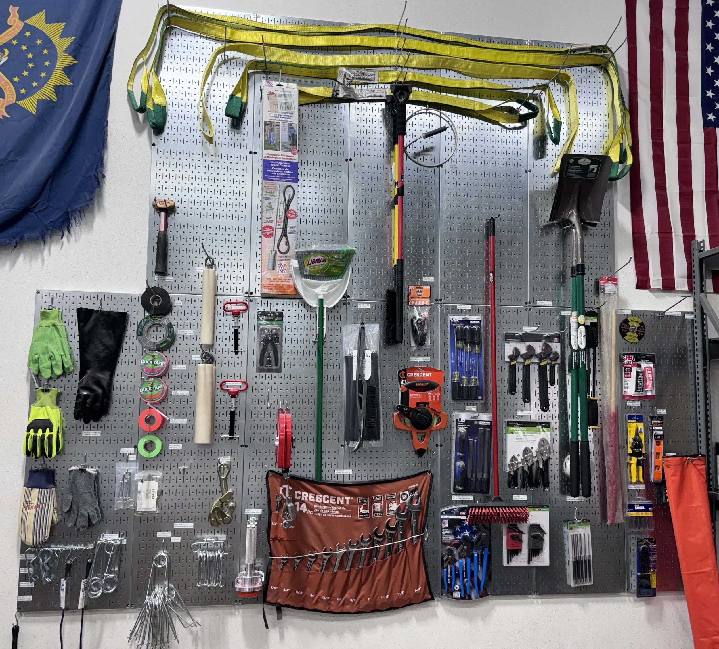 A wall display of various tools and supplies, including gloves, tape, wrenches, screwdrivers, a level, a chainsaw, and other hardware items, organized on a pegboard with flags on either side.