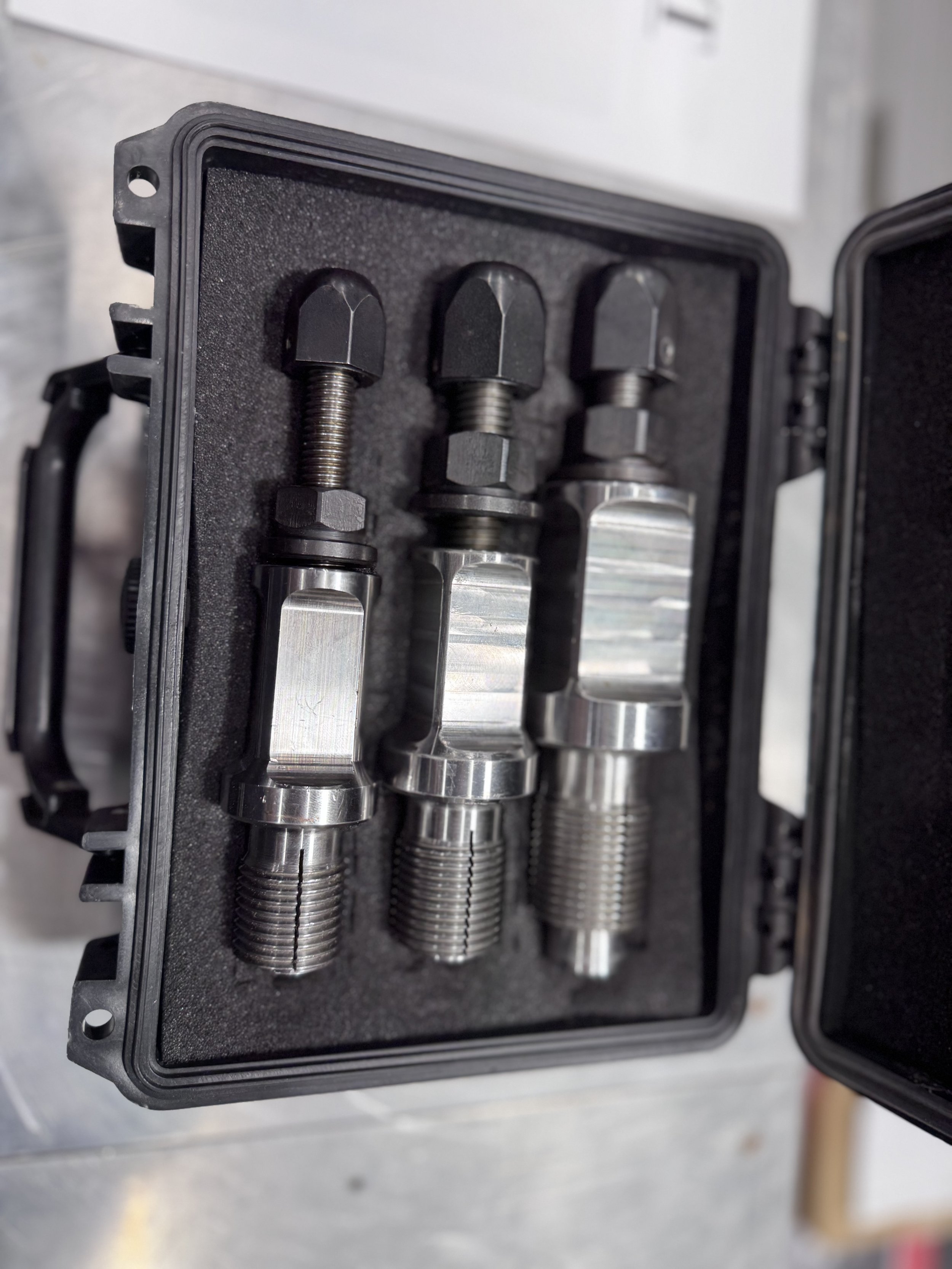 Set of three metallic engine or industrial parts with threaded sections, held in a foam-lined black case.