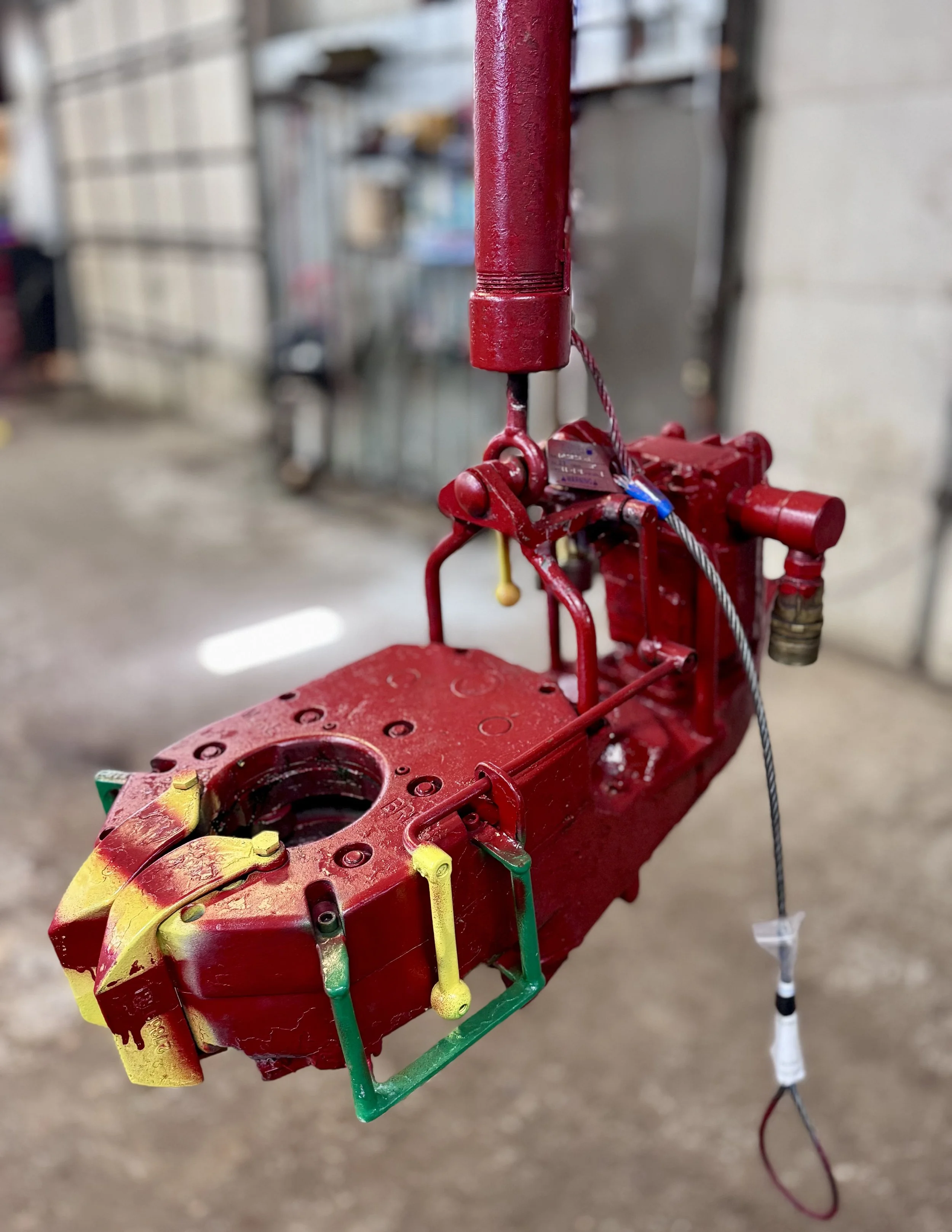 Close-up of a red mechanical device, possibly a part of a fire alarm or sprinkler system, with a hole in the center and various colored levers attached, hanging in an industrial or workshop setting.