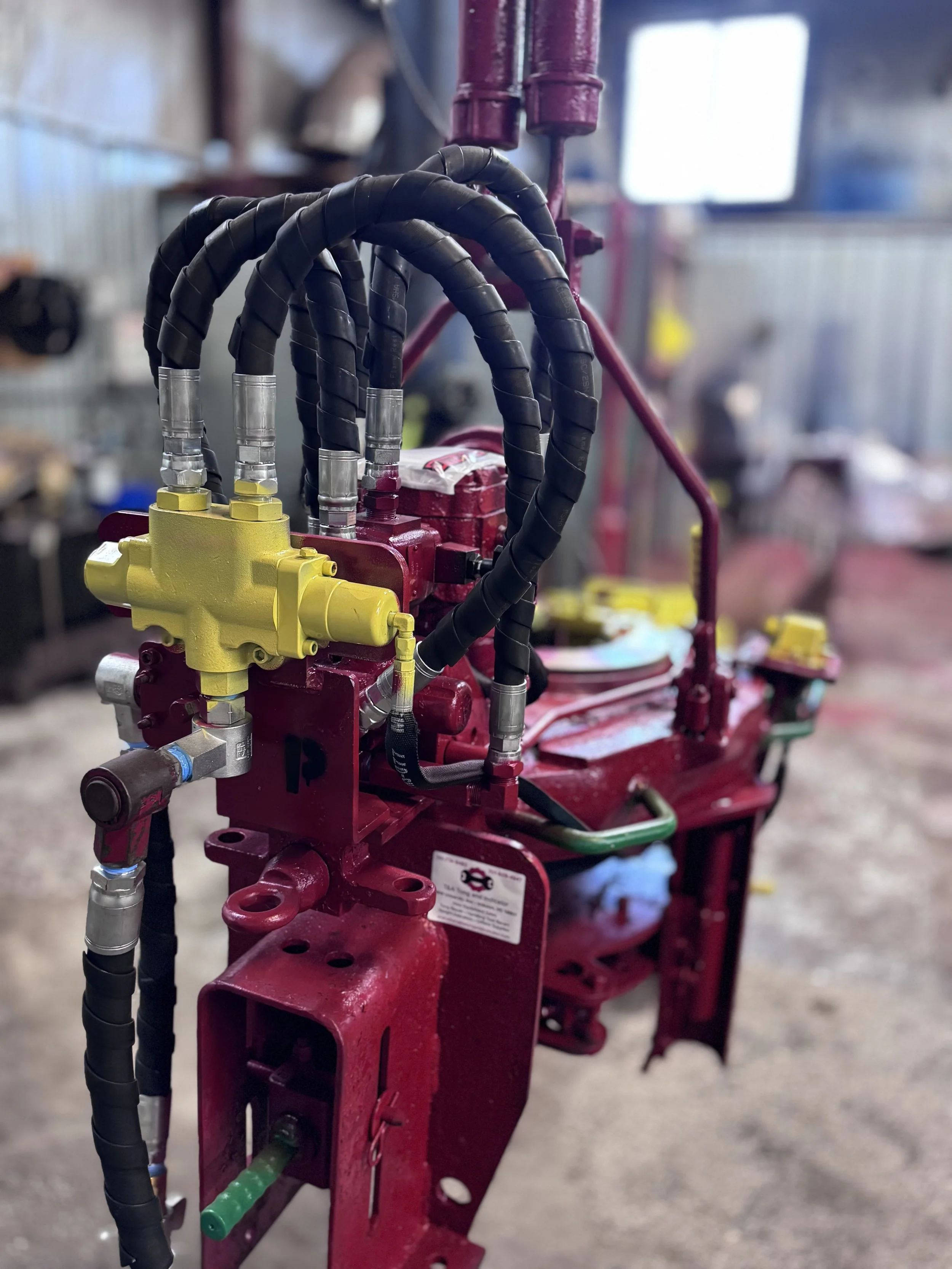 A close-up of machinery with red and yellow components, black hoses, and metallic fittings, set inside a workshop or industrial setting.