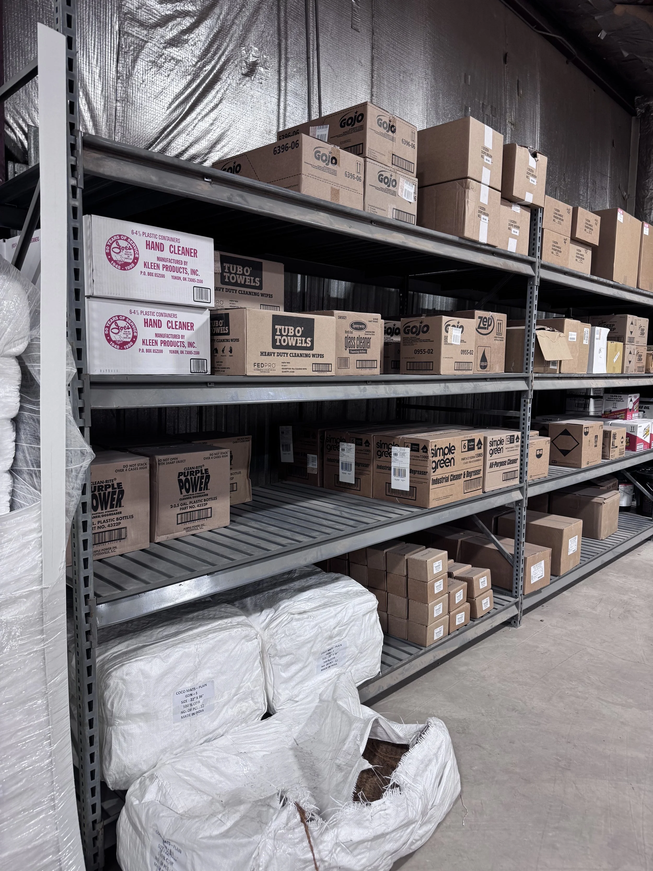 Steel shelving unit with boxes containing cleaning supplies and large bags of industrial mats in a warehouse.