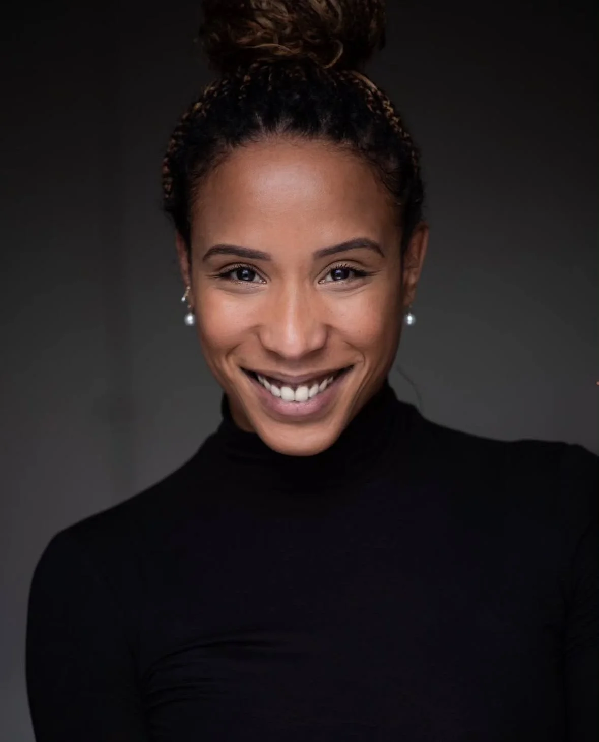 A woman with a smile, wearing a black turtleneck and pearl earrings, against a dark background.