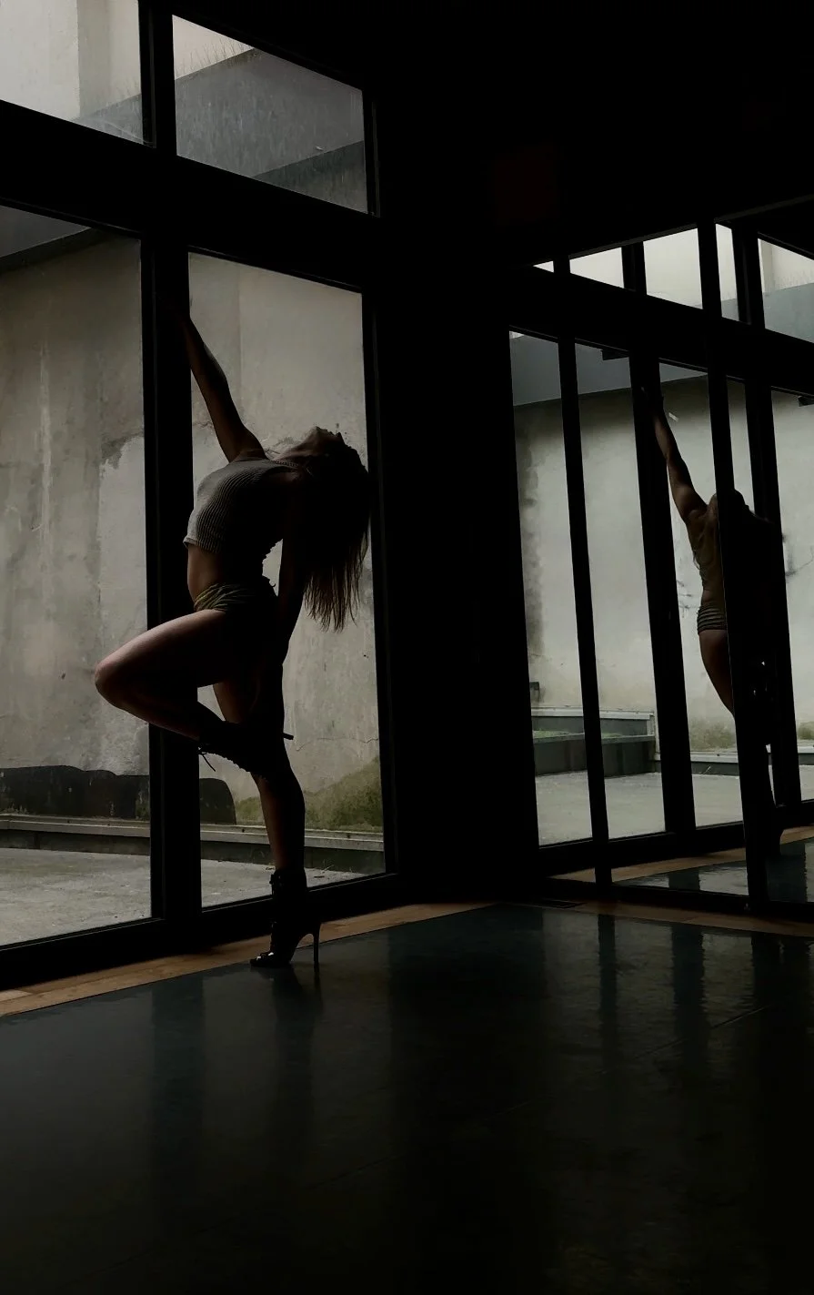 Silhouette of a woman in high heels doing a yoga pose by large glass windows in a dimly lit space, with her reflection visible in the glass.