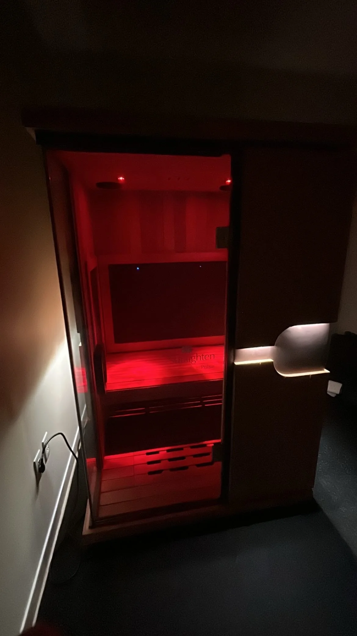 A wooden infrared sauna with red interior lighting, located in a dimly lit room.