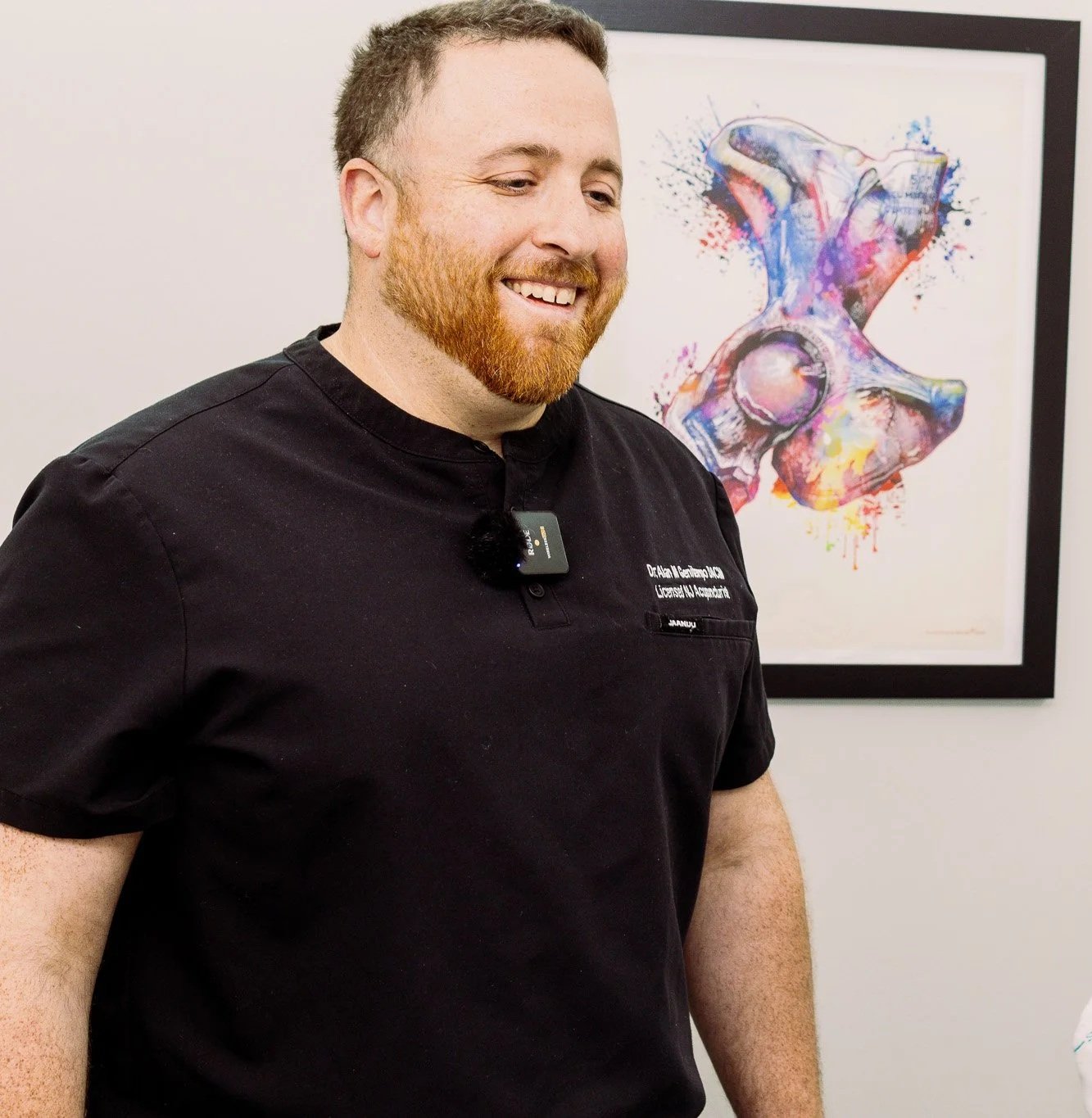 A smiling man with a beard, wearing a black shirt, standing in front of a colorful abstract artwork of a human pelvis.