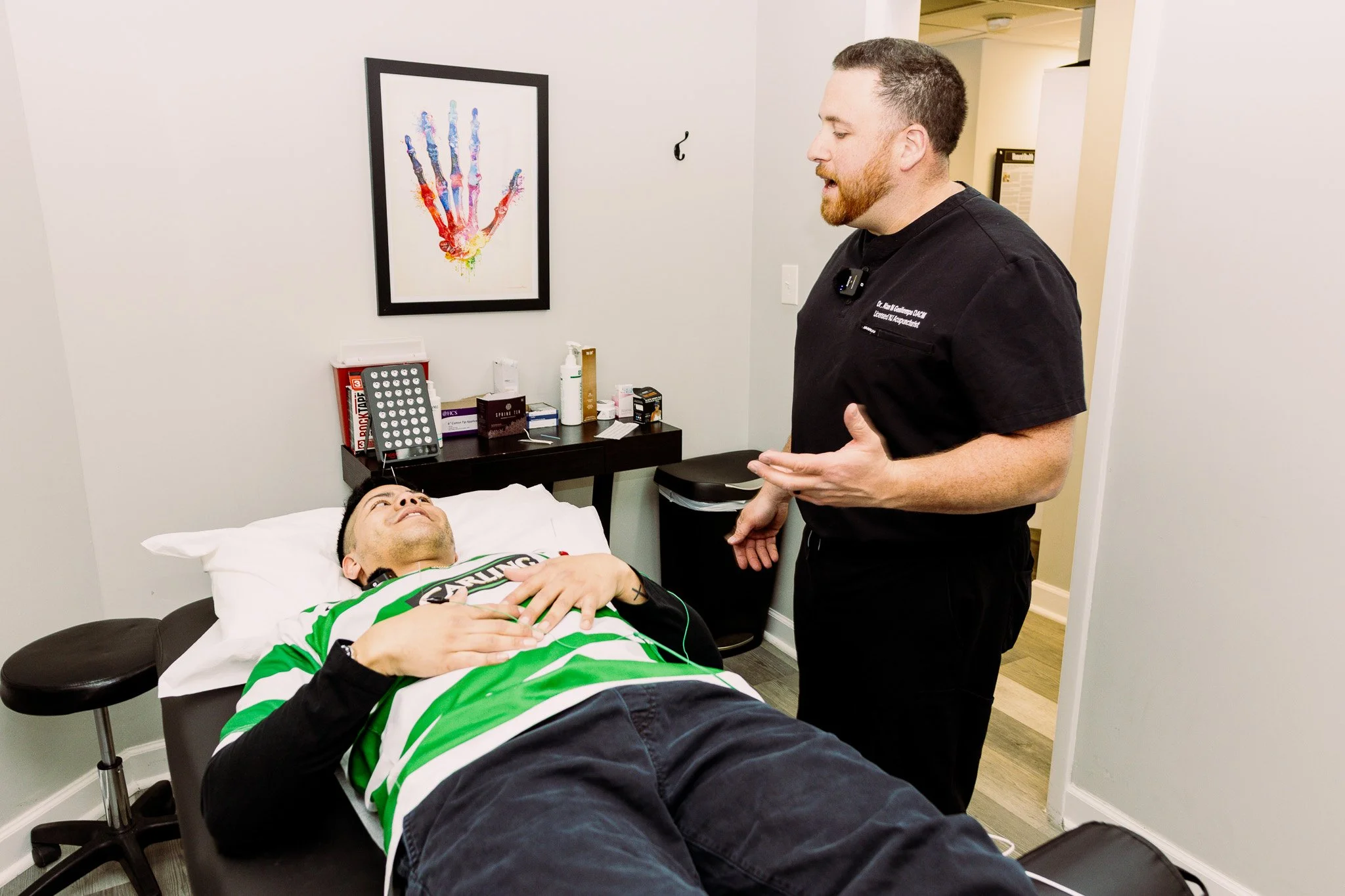 A man lying on a medical examination table, smiling, while a healthcare professional, possibly a doctor, talks to him. The man is wearing a green and black sports shirt, and the healthcare professional is dressed in black scrubs with a badge. The room has a framed colorful handprint picture on the wall and medical supplies on a nearby counter.