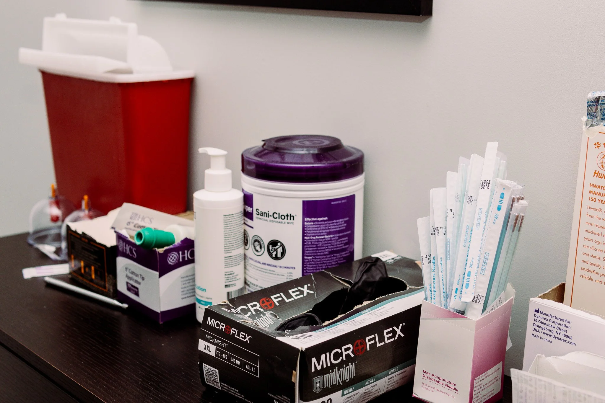 Medical supplies on a wooden table, including a red sharps container, bottles of disinfectant, boxes of medical gloves, a package of Microflex nitrile gloves, and acupuncture needles in a pink box. A white wall is in the background.