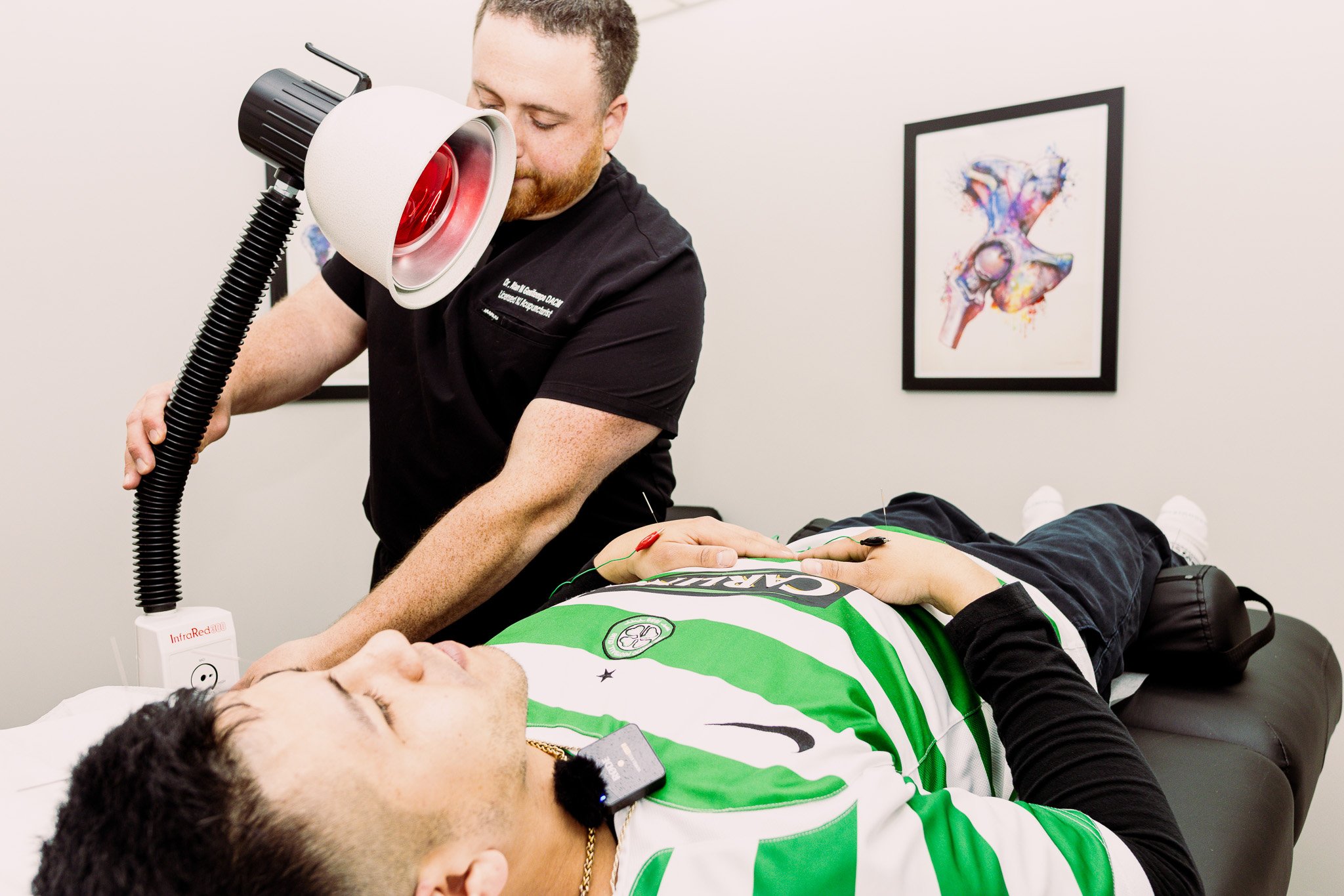 Doctor performing a medical procedure on a patient lying on a table, with a framed anatomical artwork on the wall in a medical office.