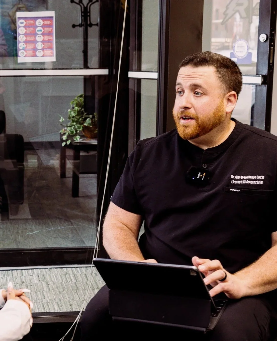 A man with a beard wearing a black shirt with medical embroidery is sitting indoors, holding a tablet. There is a glass door behind him and a poster on it, a potted plant can be seen through the glass.
