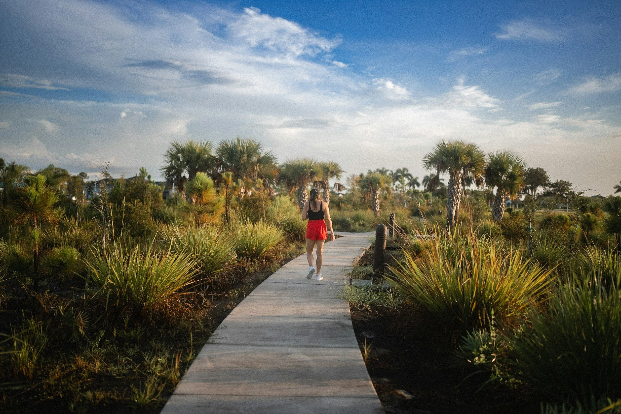 A woman in a black top and red shorts on a boardwalk thorugh tropical greens on both sides on a sunny day with light clouds