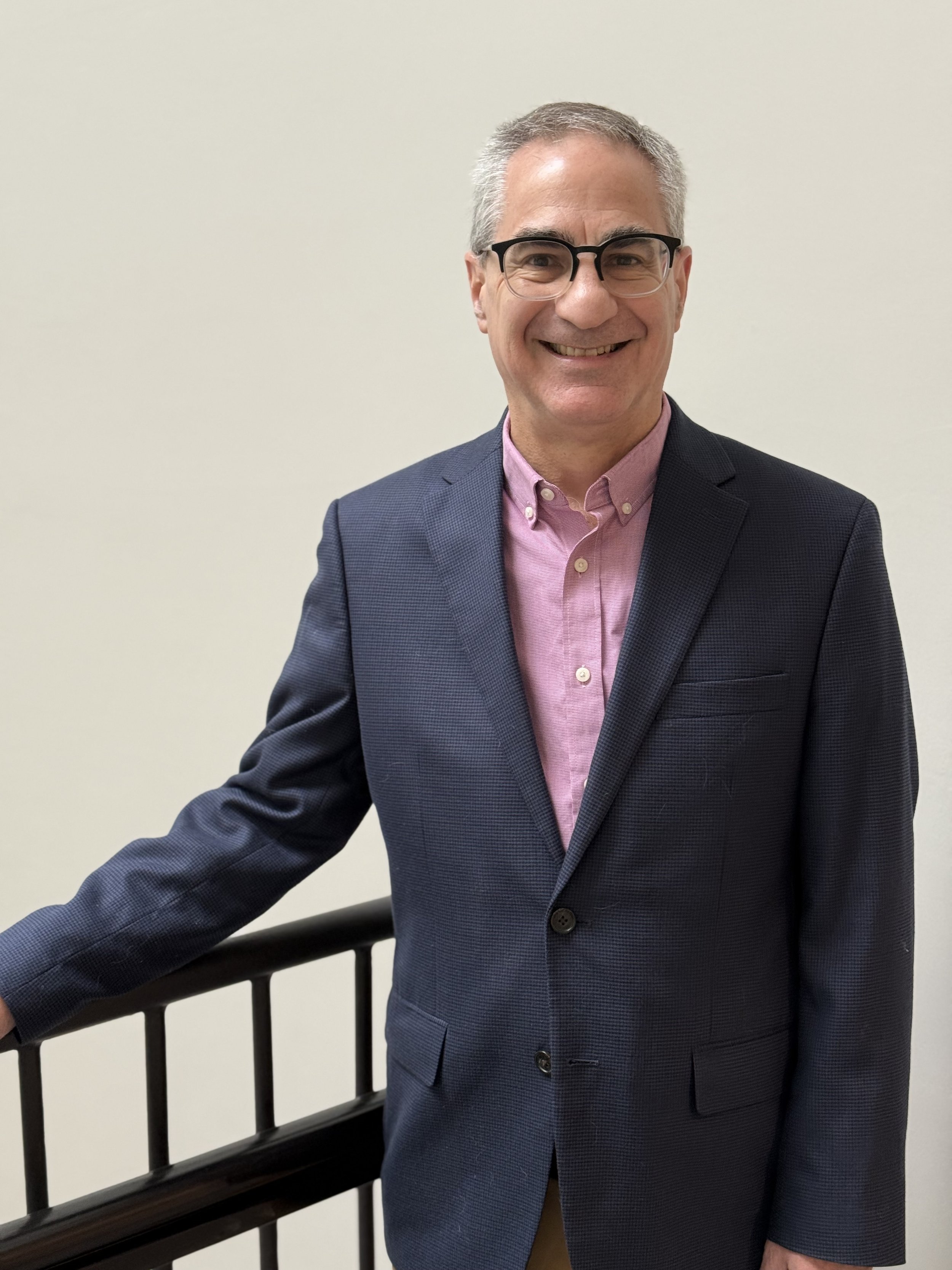 Smiling man in glasses wearing a pink button-up shirt and dark blazer standing next to a black handrail against a plain white wall.