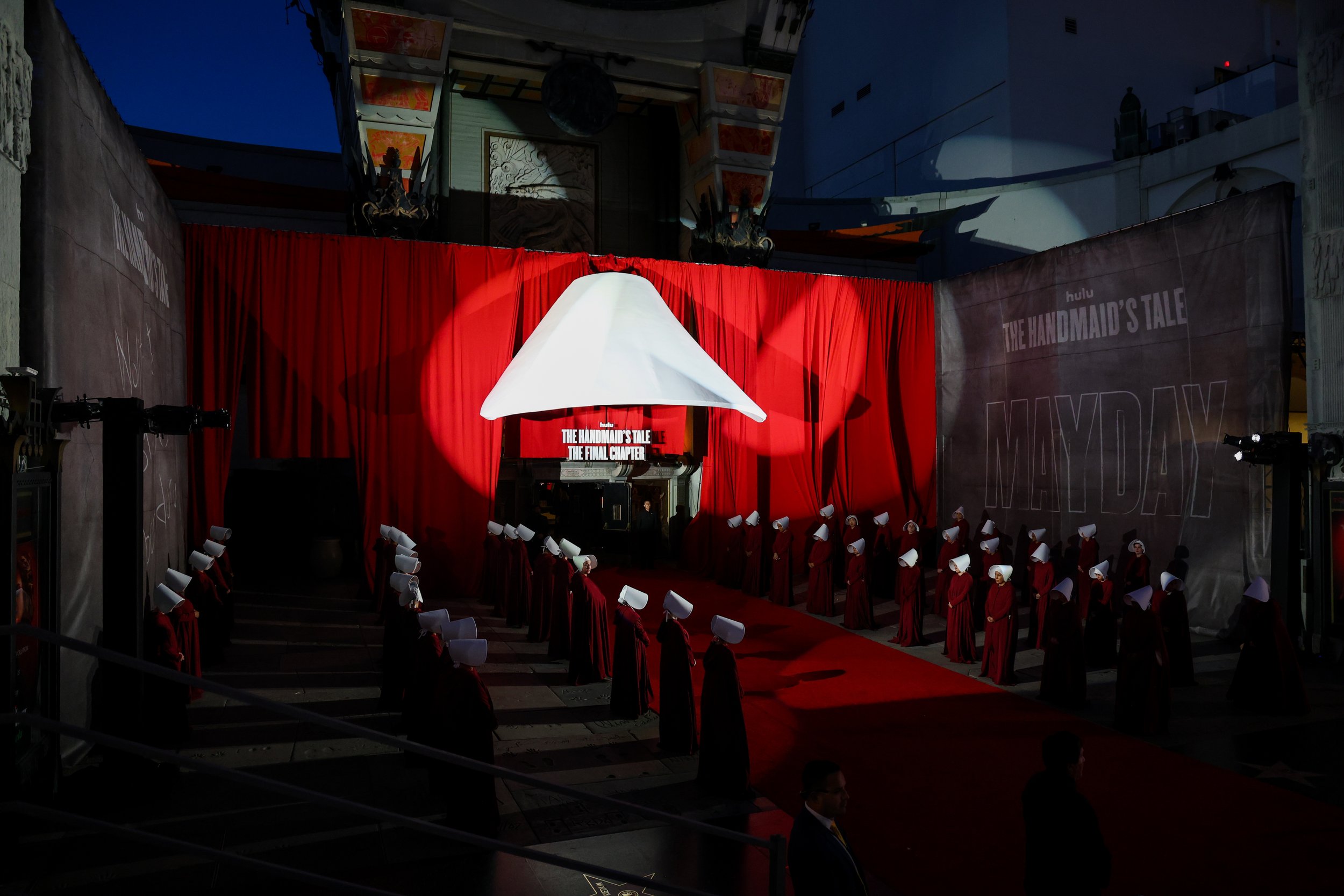 Red carpet event with people dressed in red robes and white head coverings, set against a backdrop with the logo of Hulu and the title 'The Handmaid's Tale: The Final Chapter' displayed on a screen and banners.