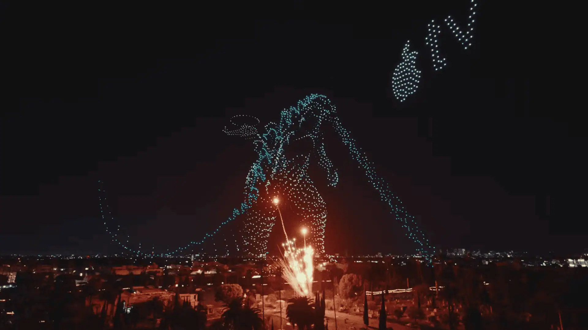 Nighttime fireworks display in the sky featuring an image of a dragon formed by lights, with fireworks at the dragon's feet.