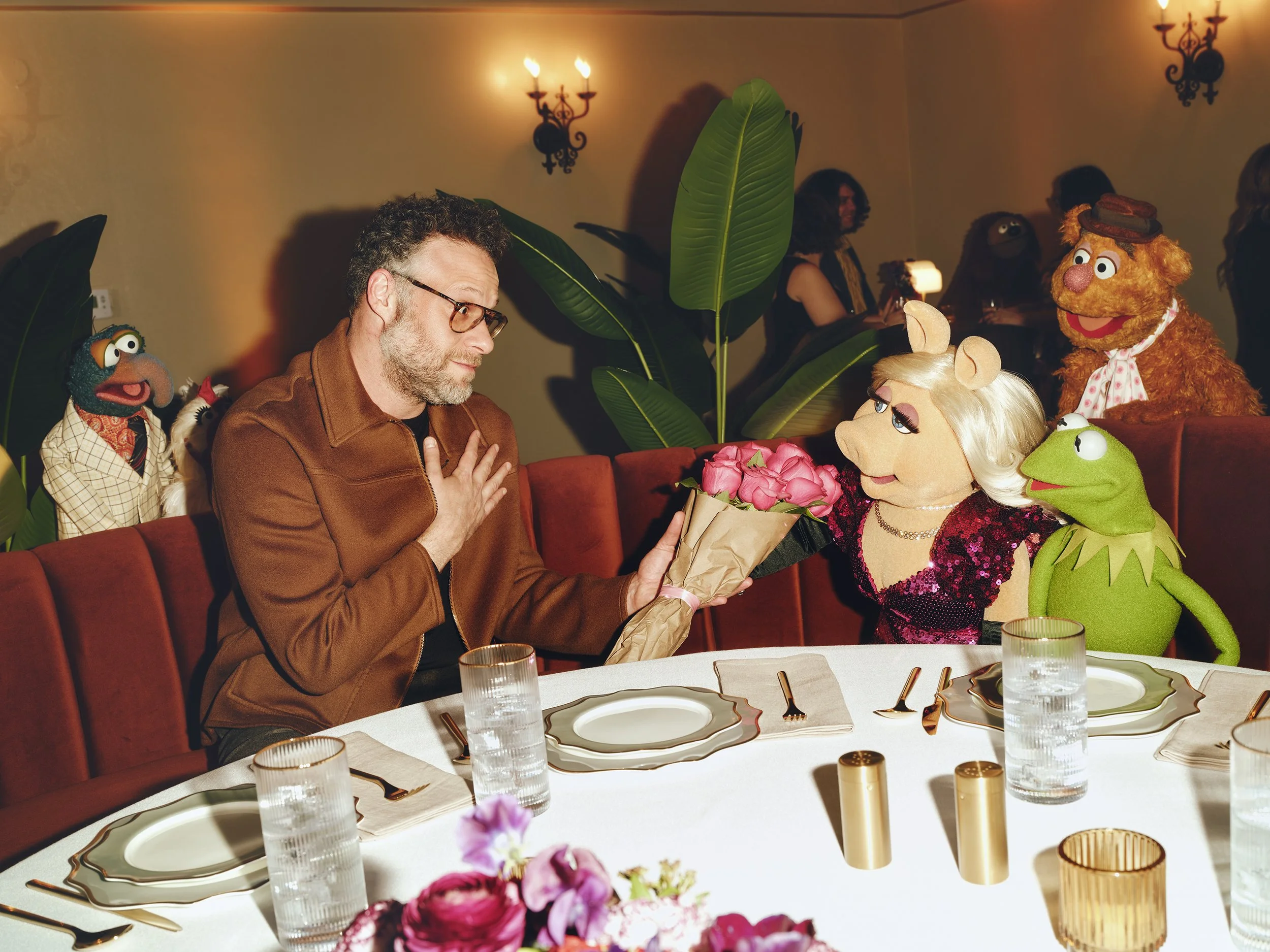 A man with glasses and a brown jacket sits at a dinner table, receiving a bouquet of pink roses from a woman dressed as Miss Piggy. The woman has blonde hair, mouse ears, and a sparkling dress. The setting is a decorated banquet with large green plants, puppets, and elegant tableware, including white plates, glasses, and candles, in a warm, dimly lit room with wall sconces.