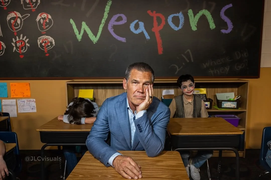 A man sitting at a desk in a classroom with a bored or tired expression, resting his head on his hand, while two children sit behind him. The classroom has a chalkboard with colorful writing that says 'Weapons' and drawings of people with question marks above their heads.