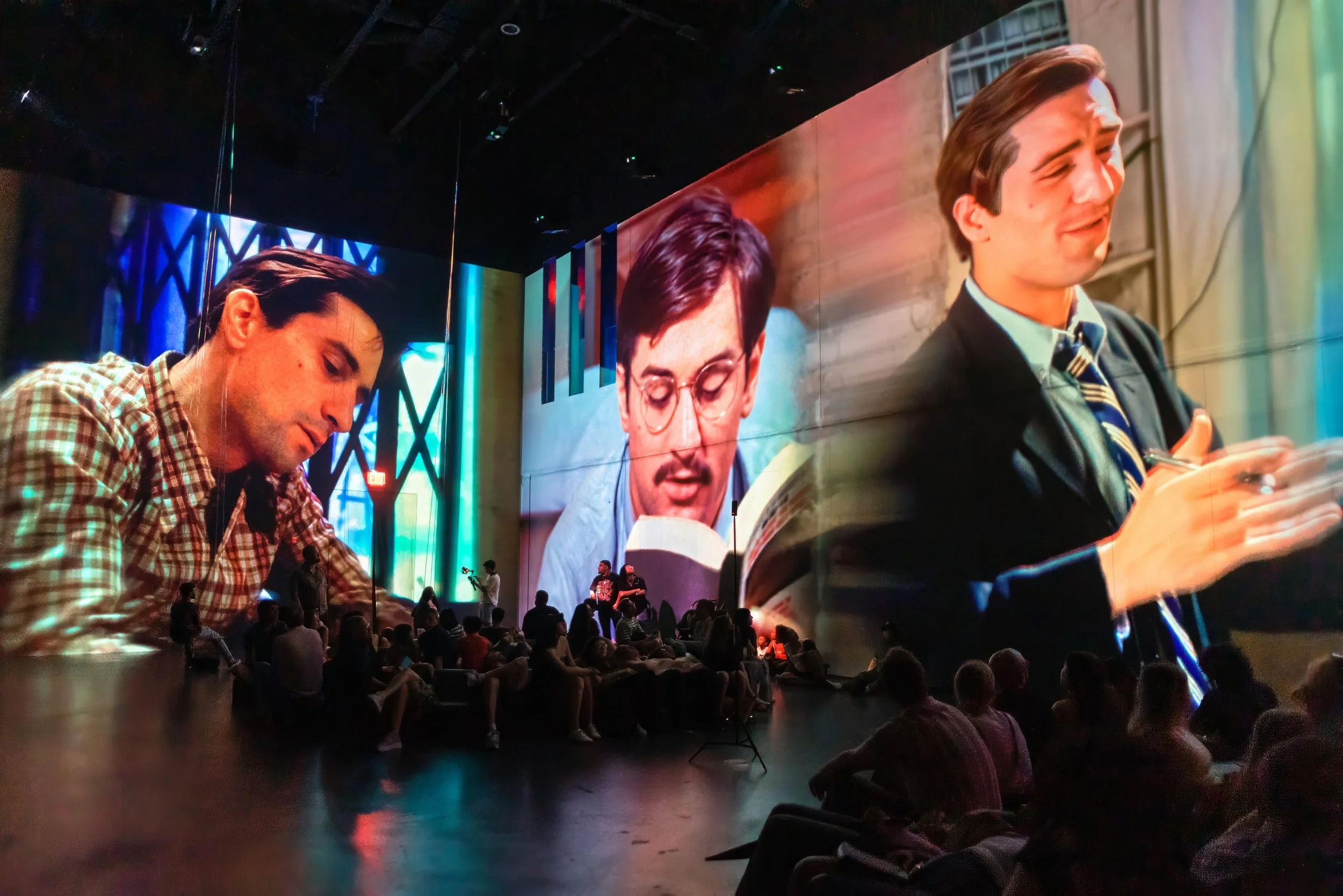 A large screen displays images of three people, with one woman and two men, in a dimly lit auditorium or conference hall. There are many people seated and standing, watching the presentation.