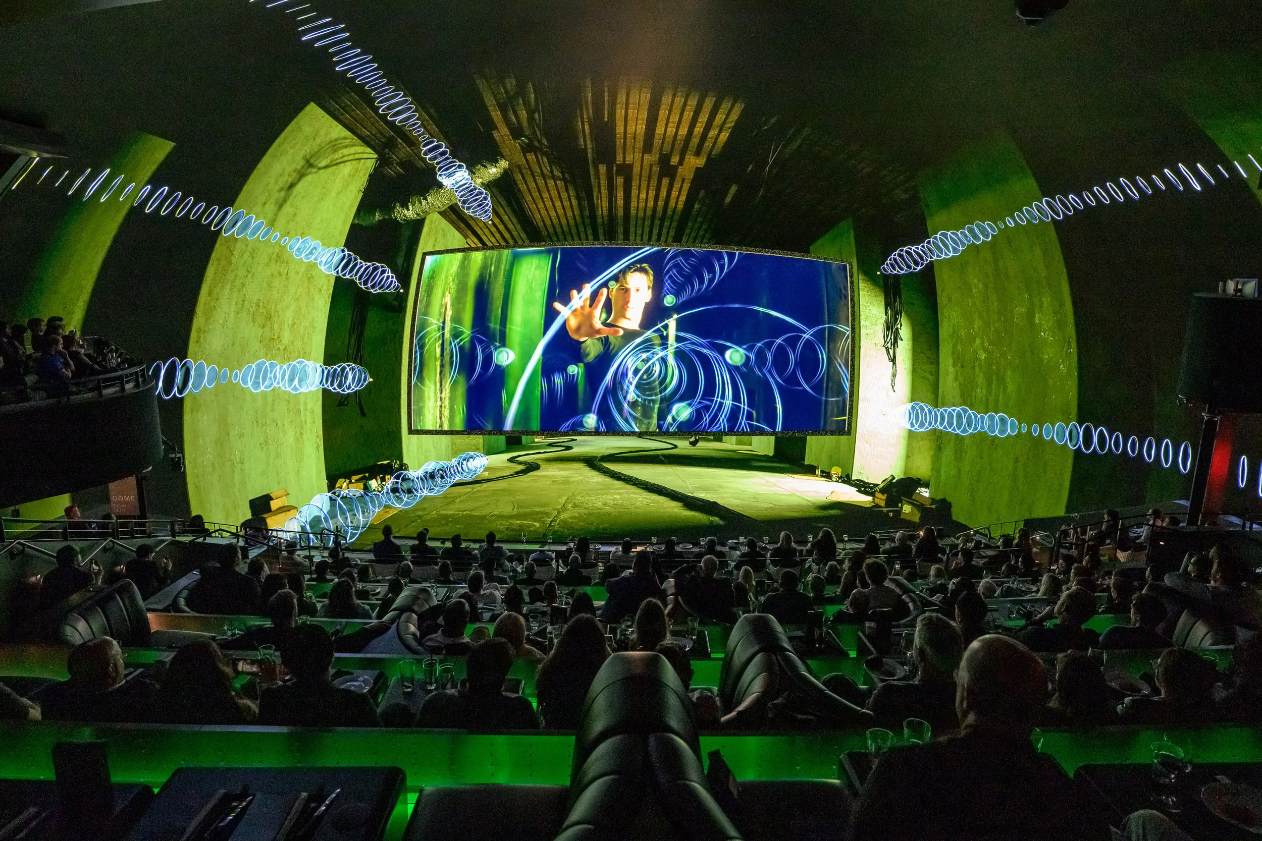 An indoor performance with an audience, featuring a large screen displaying a person reaching out with their hand, surrounded by energetic light effects and waves.