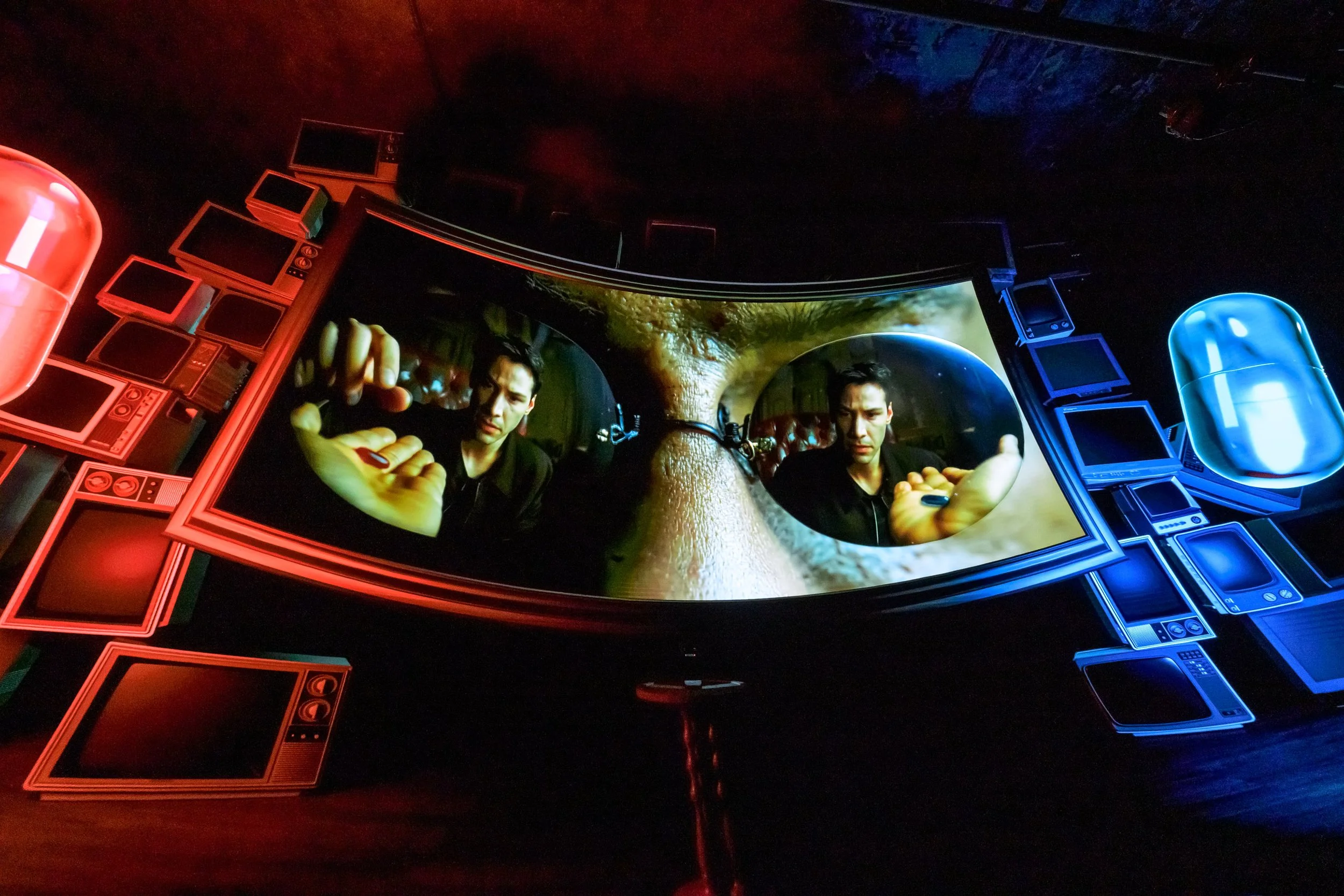 Futuristic digital artwork depicting two male figures reflected in curved screens, with vibrant red and blue lighting, surrounded by computer monitors and electronic components.