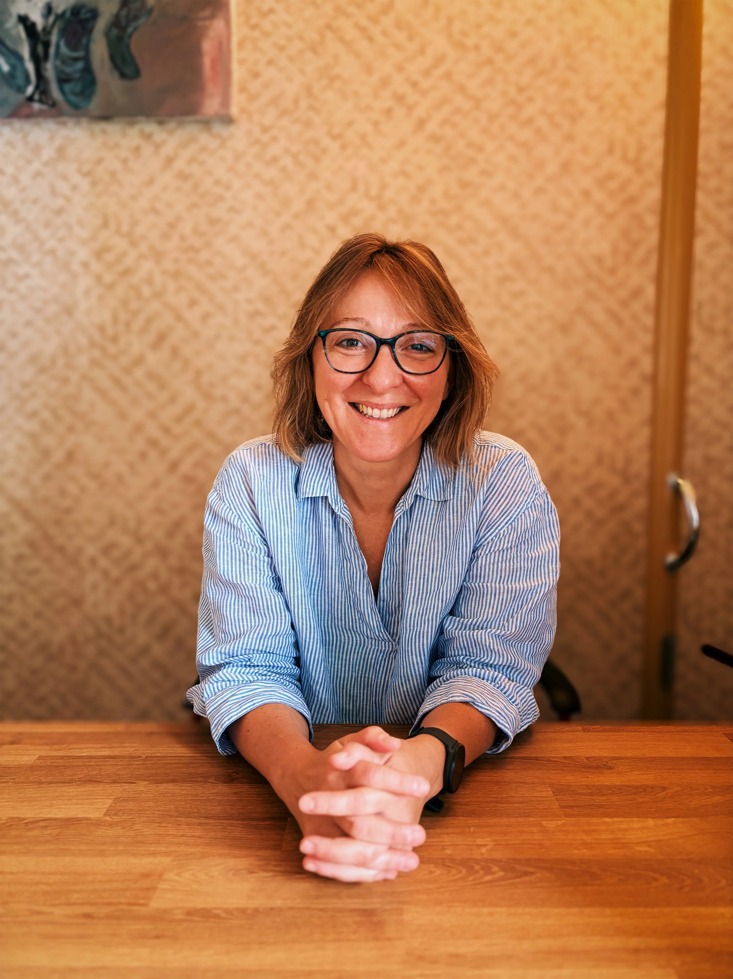 Une femme souriante avec des lunettes, portant une chemise à rayures bleues, assise à une table en bois, avec un mur beige en arrière-plan.