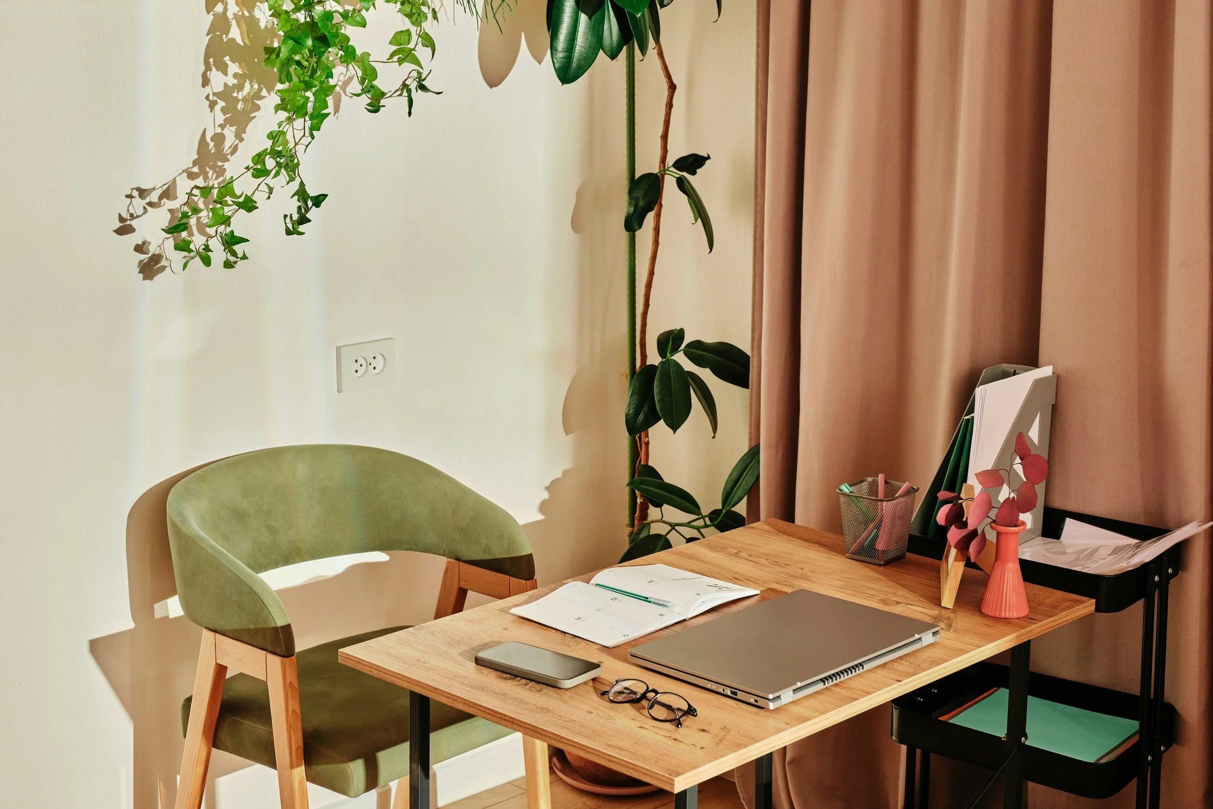 Un bureau en bois avec une chaise verte, un ordinateur portable, un carnet ouvert, une paire de lunettes, et un smartphone. Il y a des plantes en arrière-plan et des dossiers ou livres dans un porte-revues. La pièce est éclairée et a des rideaux roses clairs.