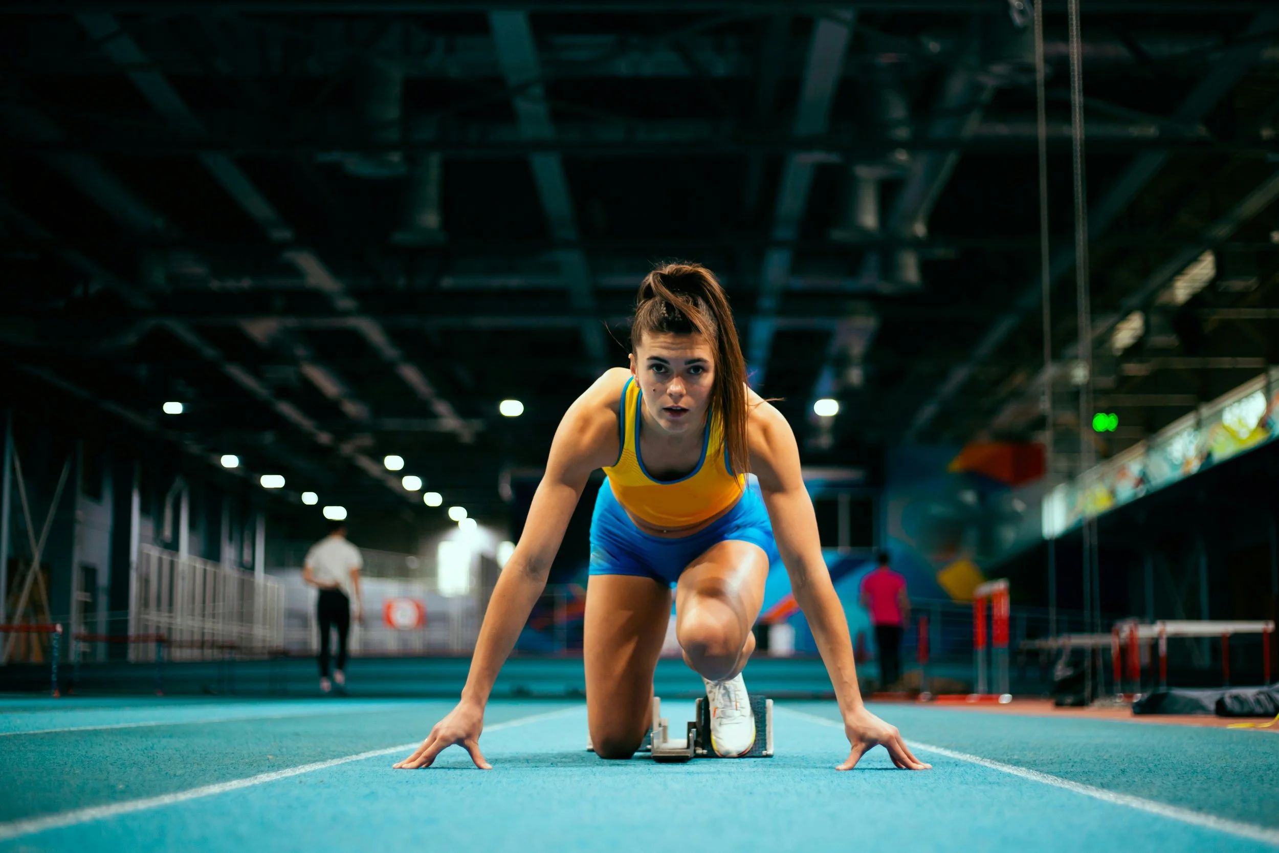 Une athlète féminine en position de départ sur une piste d'athlétisme intérieure, prête à sprinter, portant un maillot jaune et des shorts bleus, dans un environnement sportif avec d'autres personnes en arrière-plan.