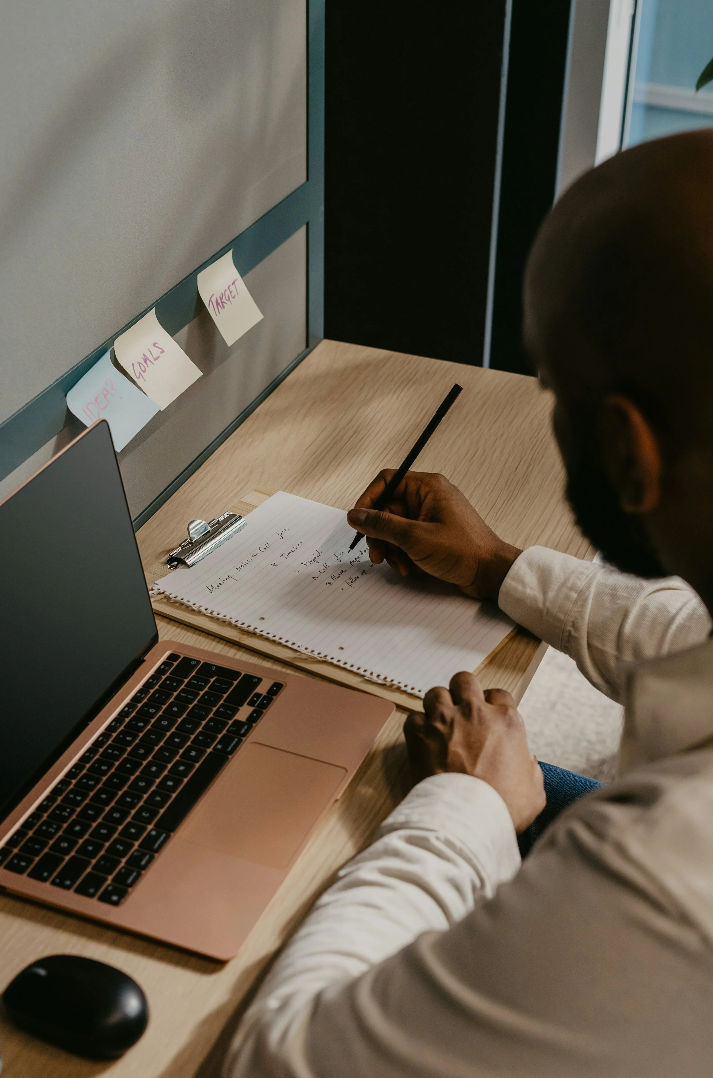 Jeune homme travaillant à un bureau avec un ordinateur portable rose, un cahier, un stylo, des post-it sur le panneau diviseur avec des mots en anglais, et une souris d'ordinateur.