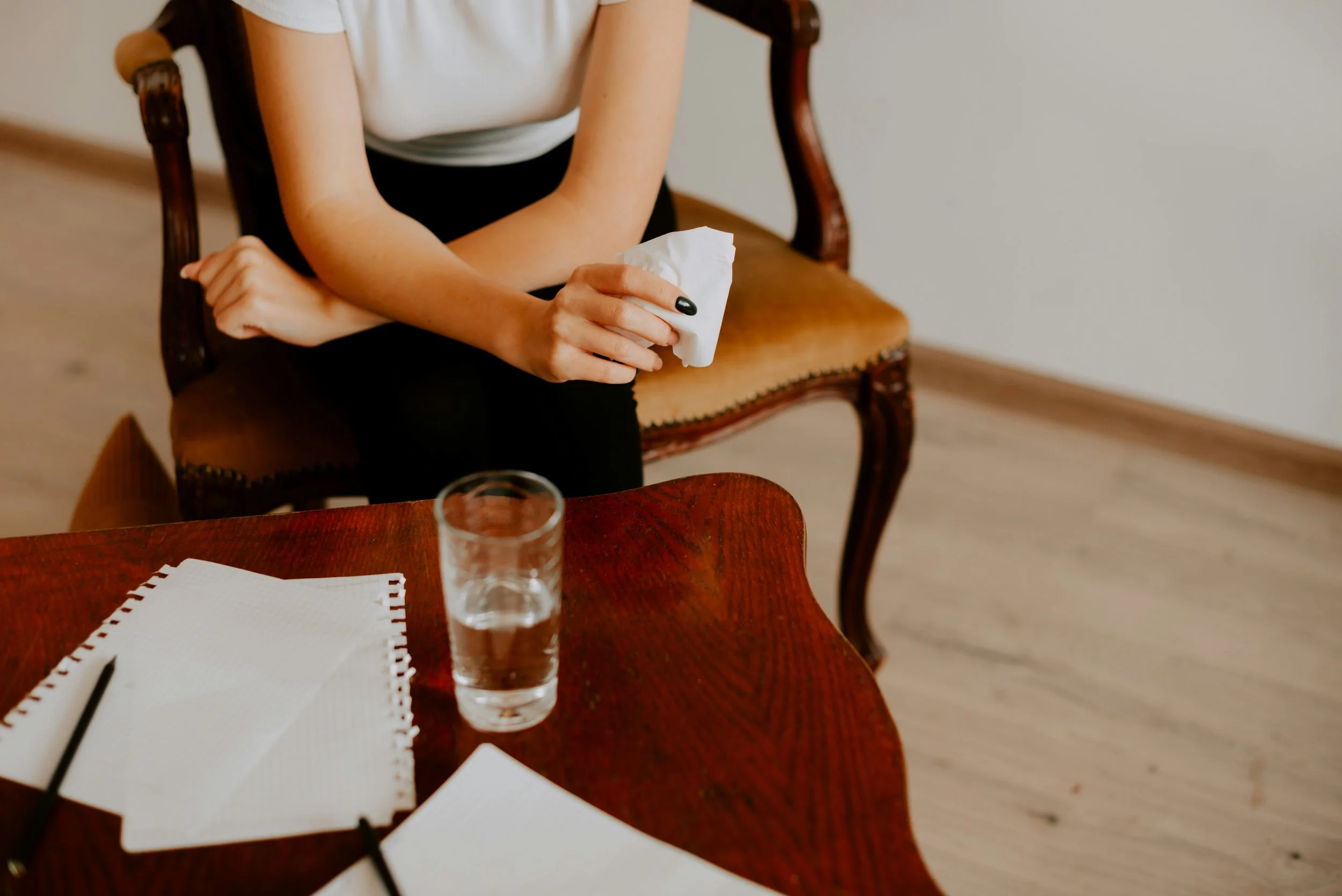 Une personne assise à une table en bois, tenant un mouchoir en papier dans la main, avec un verre d'eau devant elle. Sur la table, il y a une grille, un carnet de notes et une déclaration ou une fiche blanc.