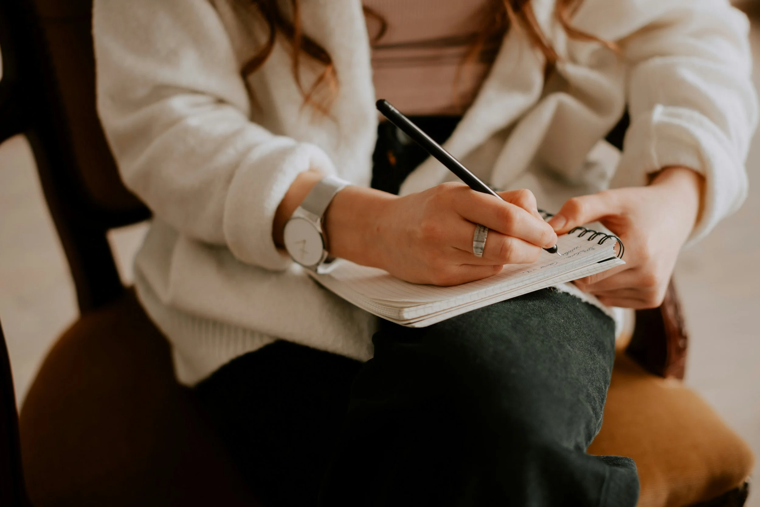 Une personne assise, écrivant dans un carnet avec un stylo, portant une montre et une bague, vêtue d'un pull blanc crème avec des manches relevées.