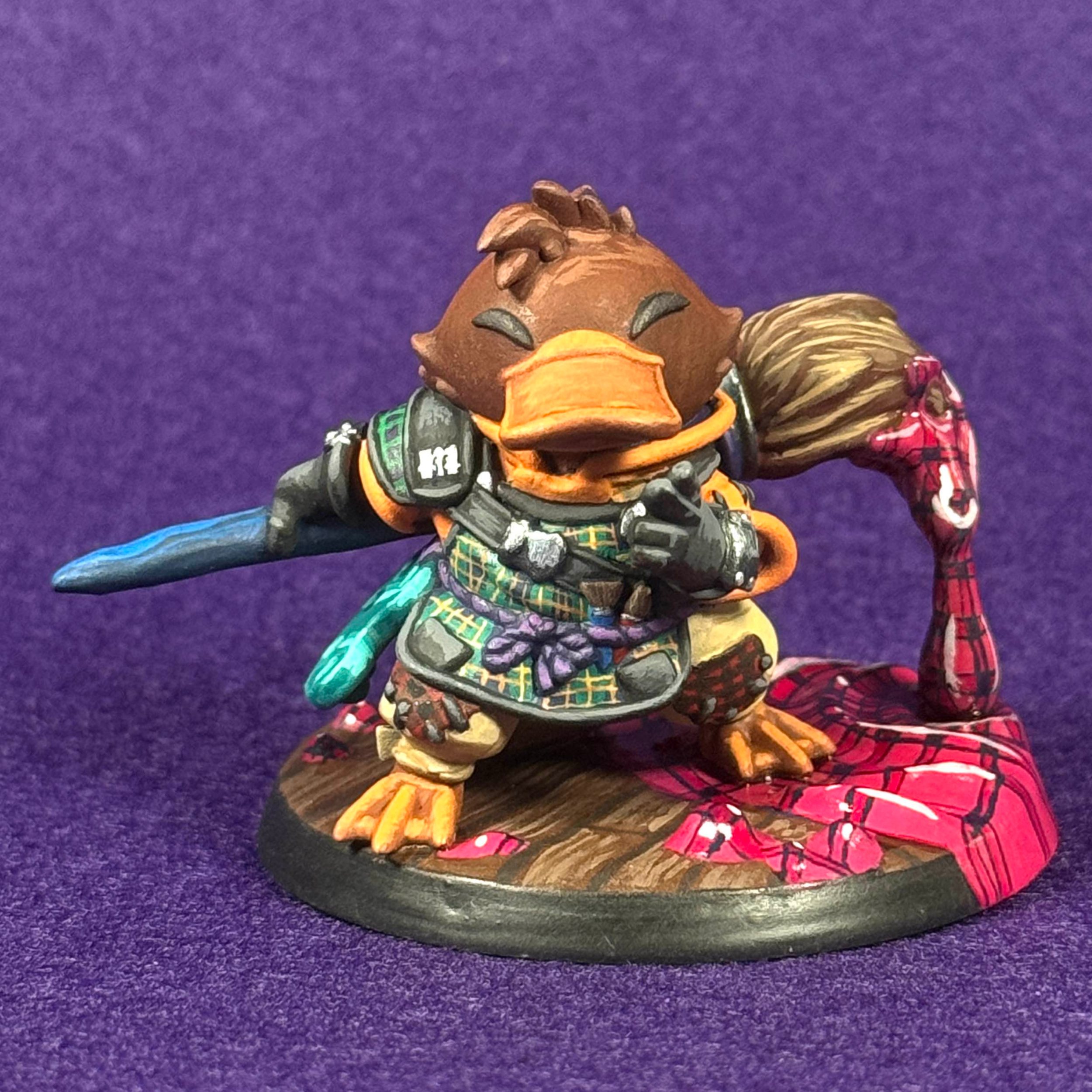 A close up photo of a 2" tall resin model of an anthropomorphic platypus weilding a gian paintbrush and wearing a painters apron. It's painting in multple different kinds and colors of plaid.