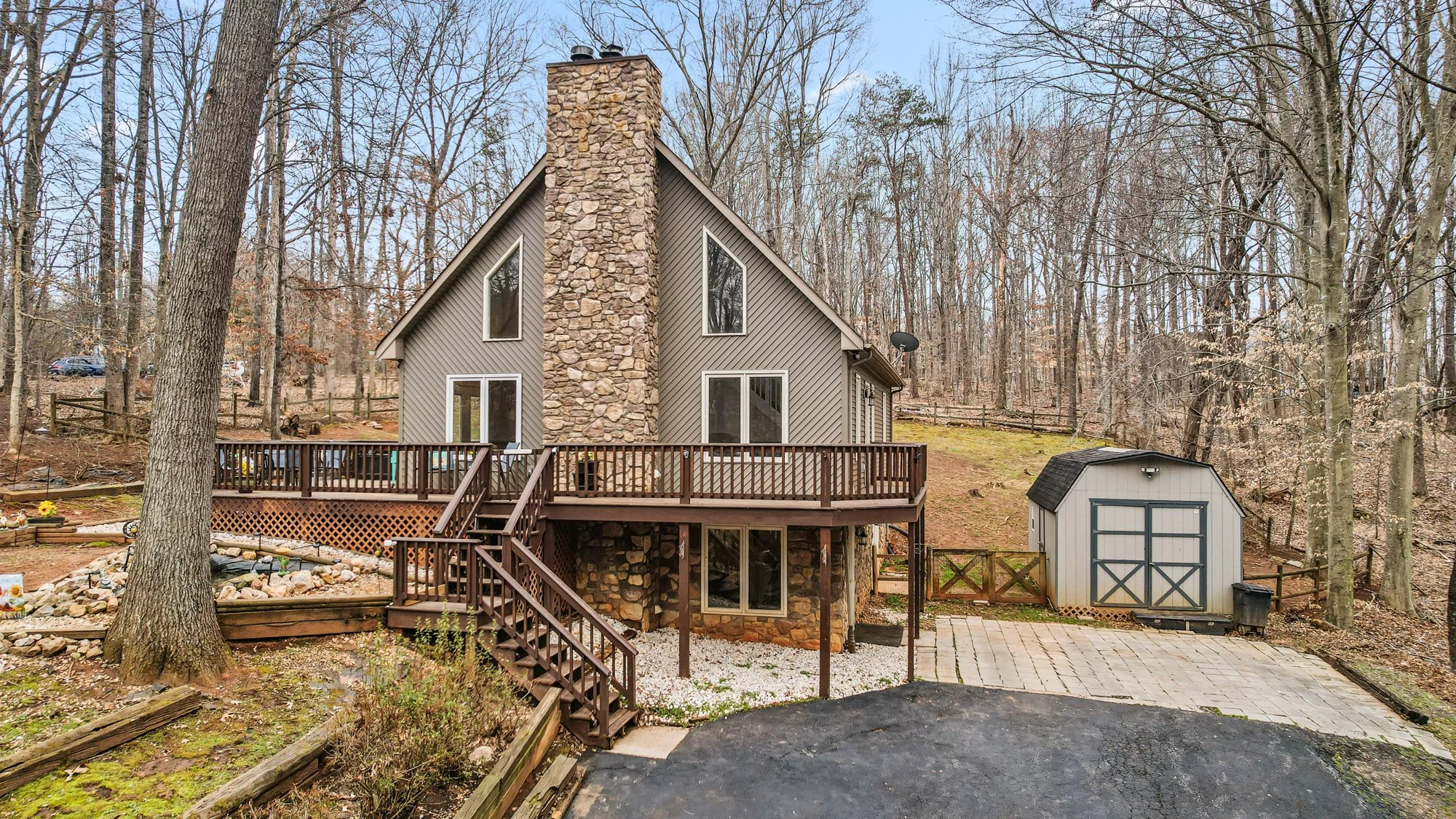 Front view of a two-story house with a stone chimney, gray siding, and large windows, surrounded by leafless trees, with a wooden deck, a paved driveway, and a small shed.