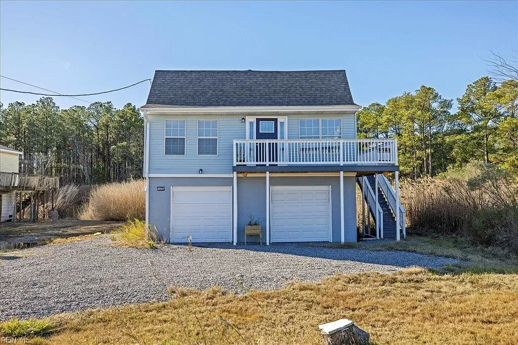 A two-story house with blue siding, white trim, a black shingled roof, and a balcony on the second floor. There are two garage doors on the ground level, and stairs on the right side leading up to the balcony. The house is surrounded by grass, trees, and a gravel driveway.