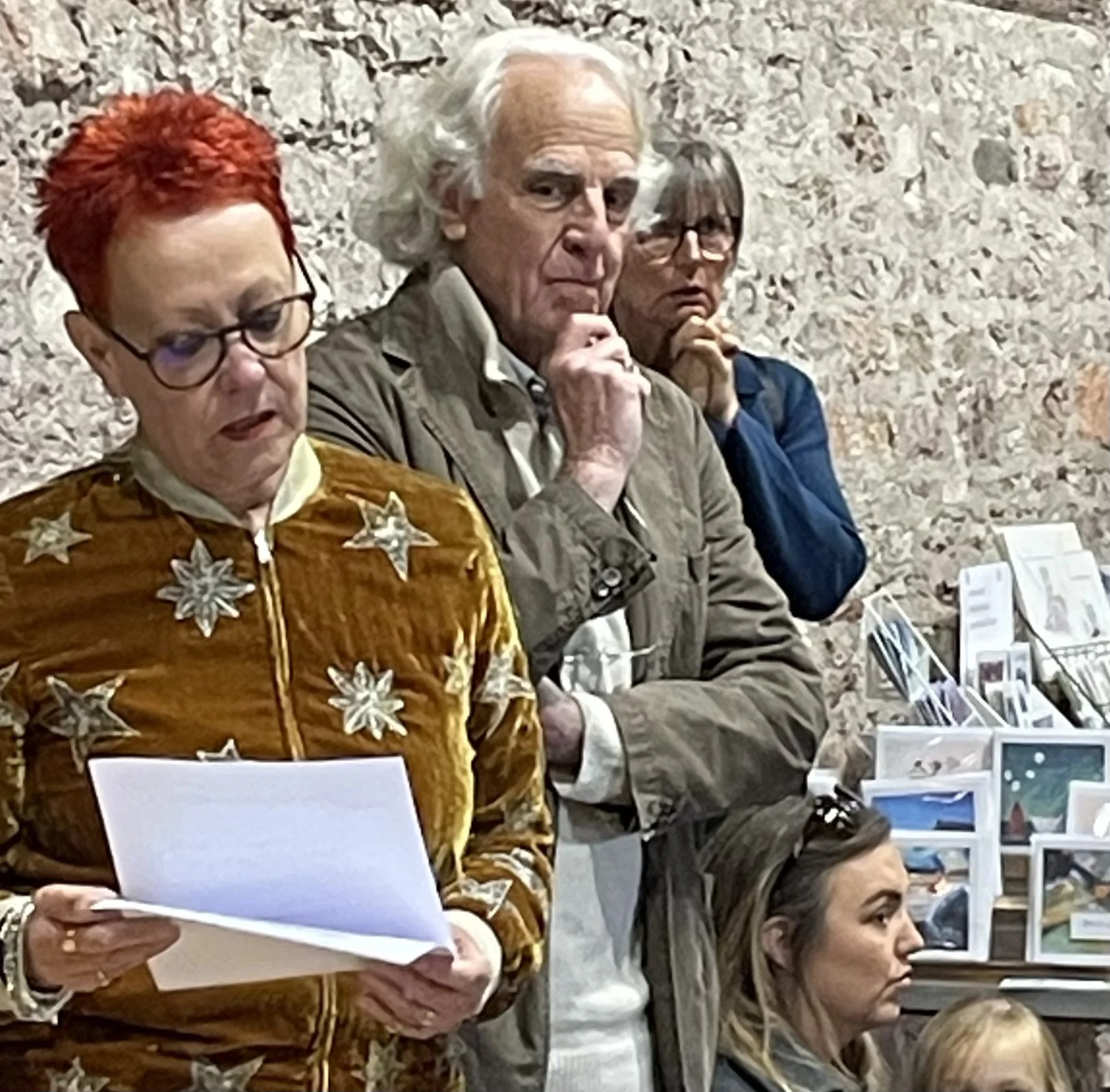 Three people sit at a table. One woman with red hair and glasses wears a brown jacket with star-shaped embellishments. A man with white hair and a beige jacket has his hand near his face, appearing thoughtful. Another woman with glasses, dark hair, and a blue shirt looks on. The background is a rough stone wall, and there are brochures or pamphlets on the table.