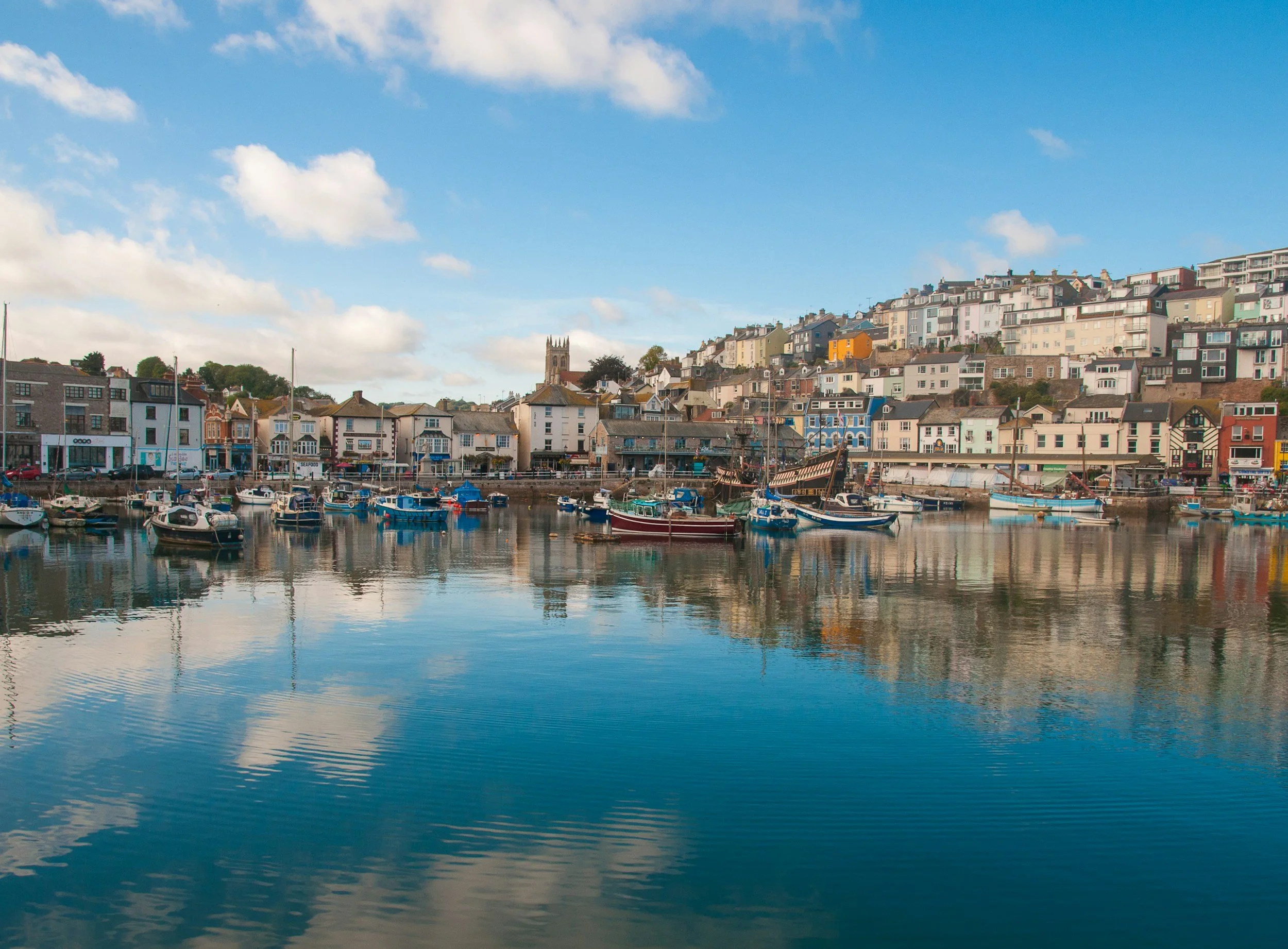 A harbor with boats docked in calm water and a hillside filled with colorful houses and a church with a tall steeple under a partly cloudy blue sky.