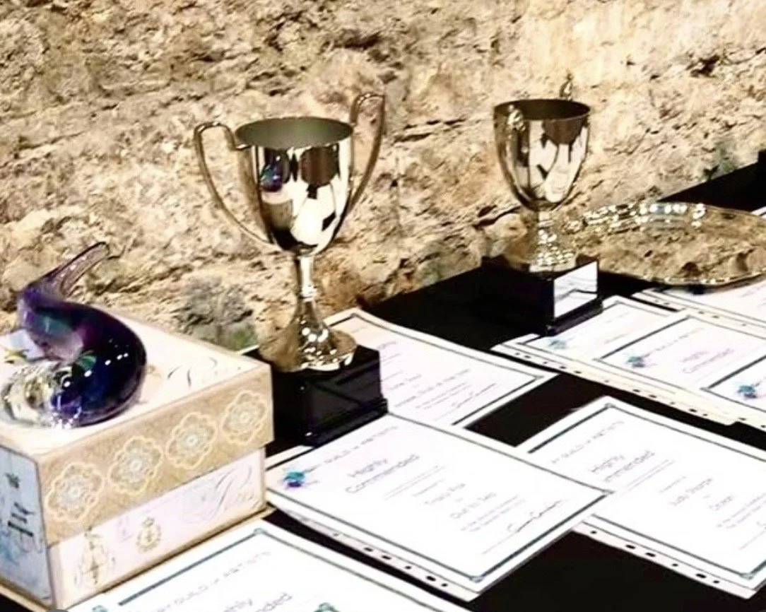 Two silver trophies, documents, a decorative glass bird, and a silver tray on a table against a stone wall.