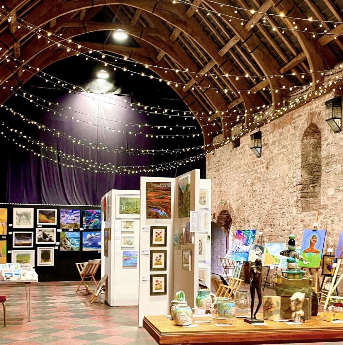 Art gallery with paintings, sculptures, and decorative items displayed on white panels and tables inside a rustic brick building with string lights hanging from the wooden ceiling.