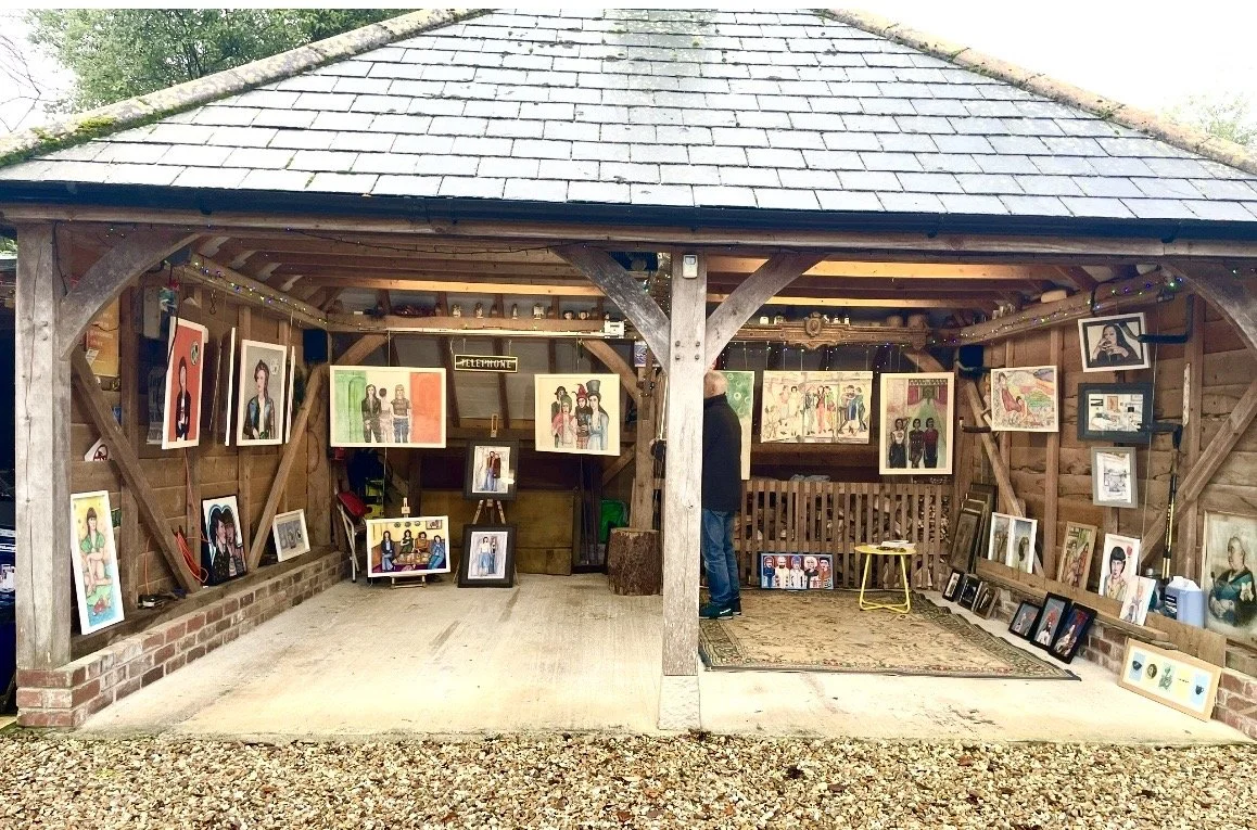 Art gallery inside a wooden shed displaying various colorful paintings of people and landscapes, with a person looking at the artwork.
