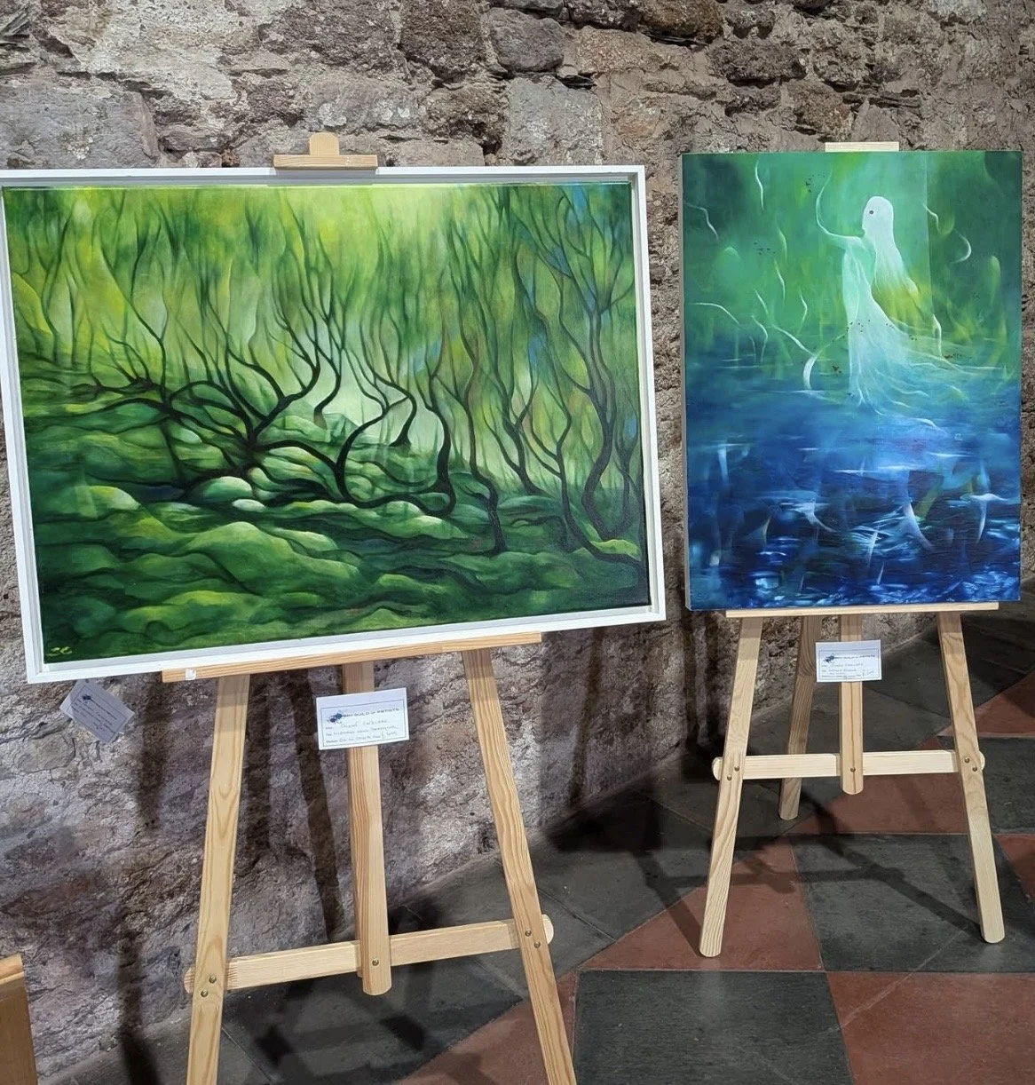 Two paintings on display easels in front of a stone wall, the left painting depicts a green forest scene with twisted trees and dense foliage, the right painting shows two glowing white swans on a blue and green water background.