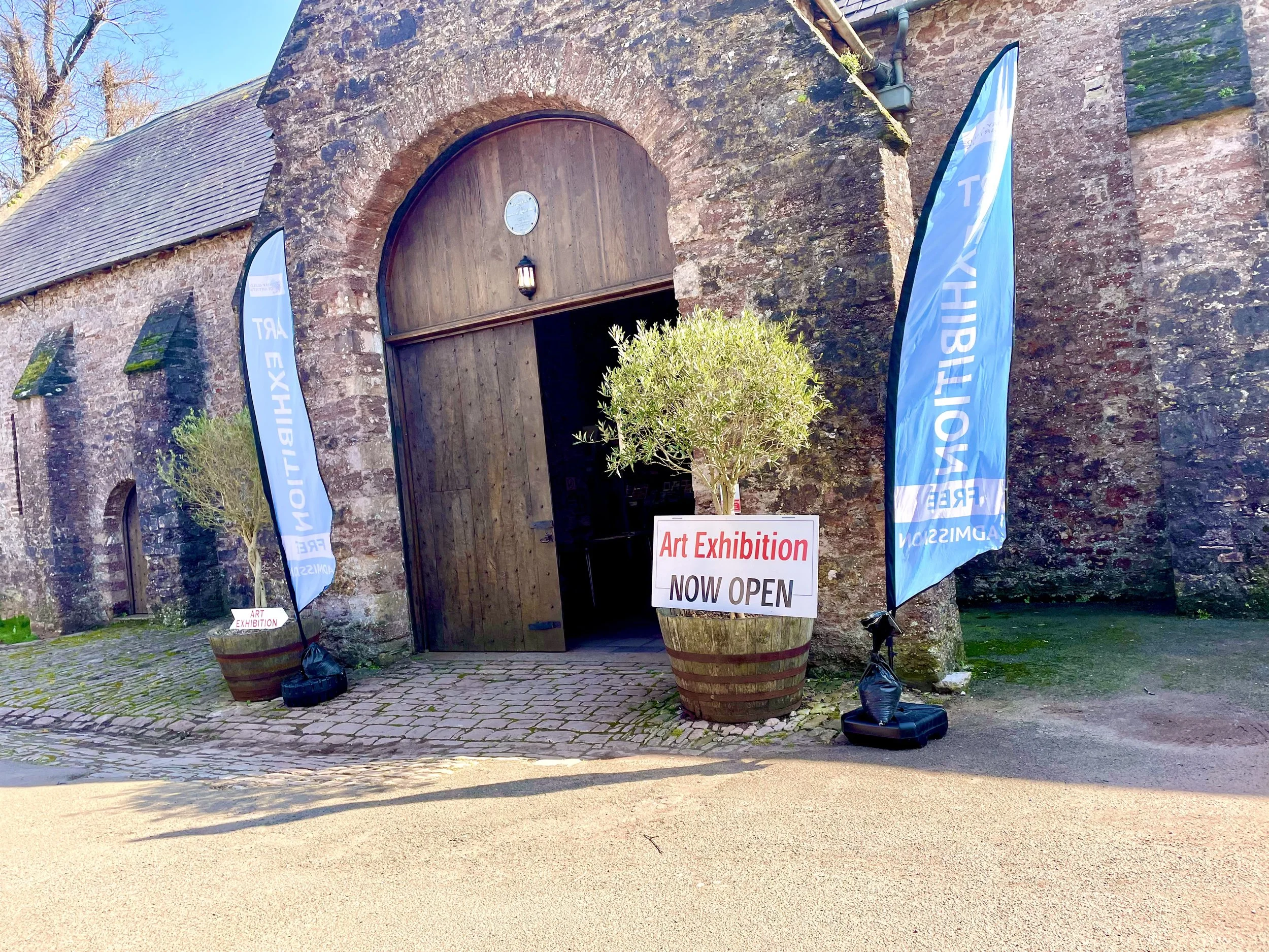Stone building with a large wooden arched door, blue flags that read 'ART EXHIBITION', a sign that says 'Art Exhibition NOW OPEN', small potted trees, and an outdoor cobblestone and sandy area.