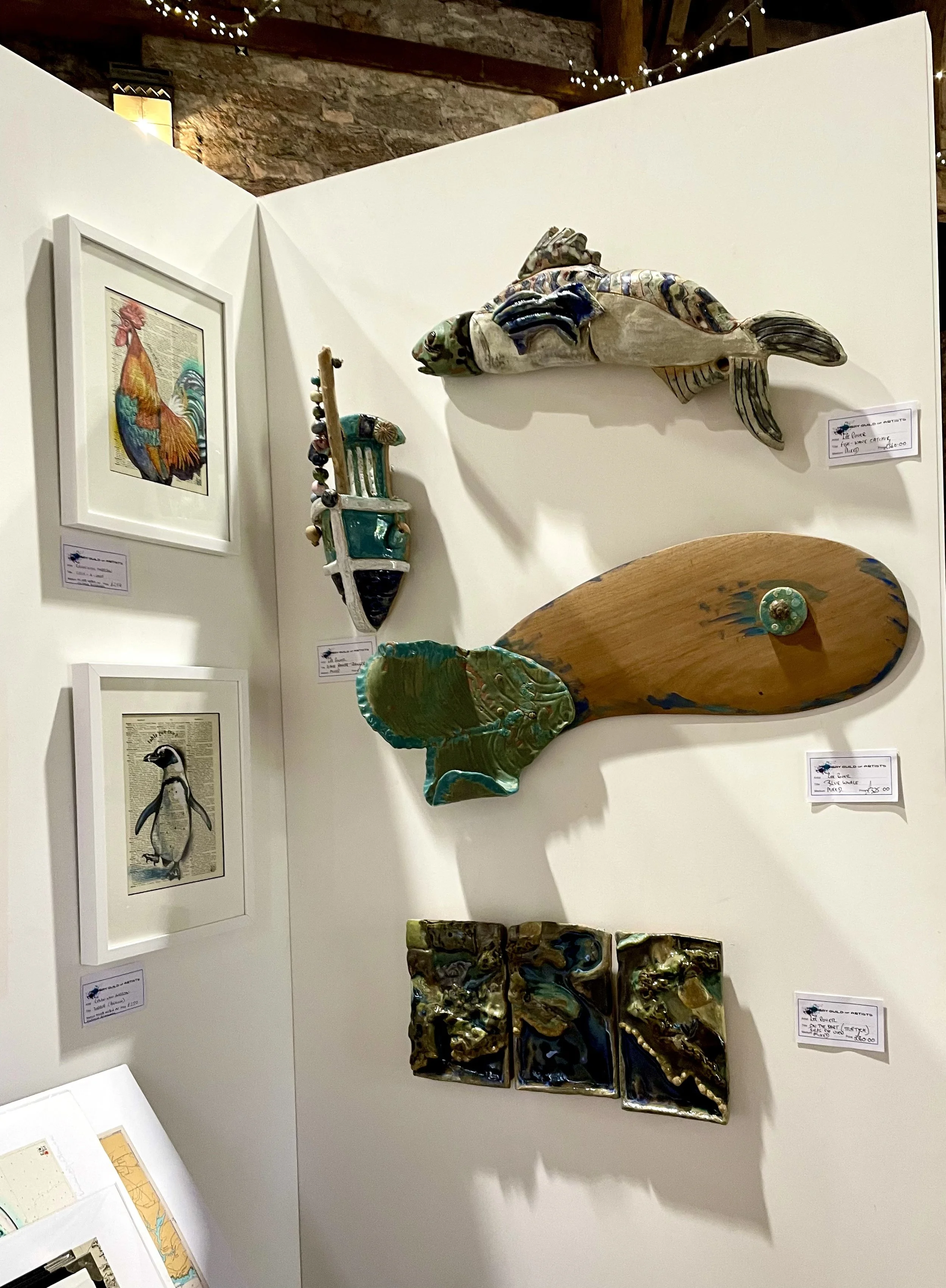 Various ceramic art pieces and framed artwork displayed on a white wall at an art exhibit, with a brick and wood ceiling and string lights in the background.