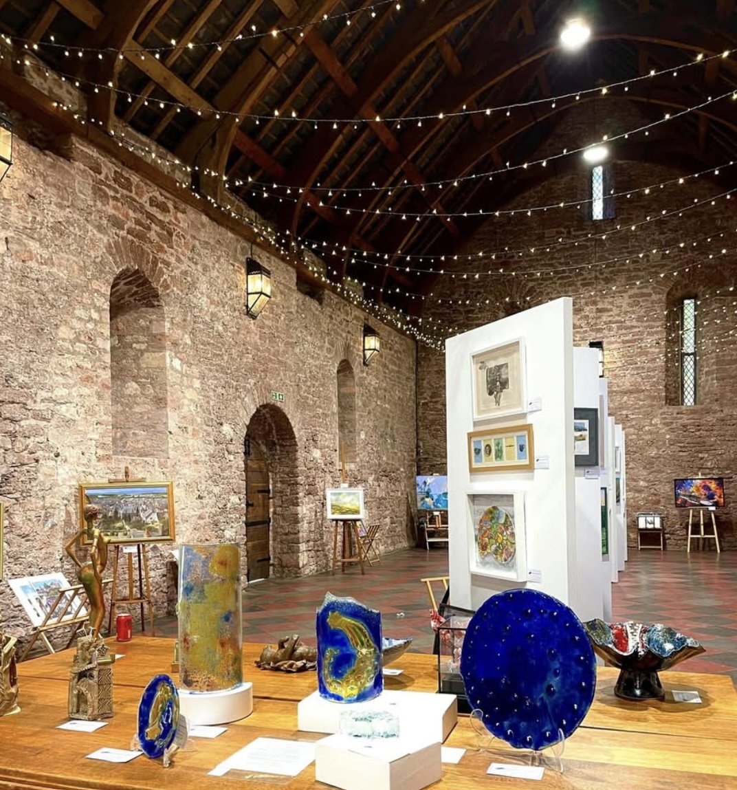 Art gallery in a large brick-walled hall with string lights on the ceiling. Various paintings and colorful glass art sculptures displayed on tables and easels.