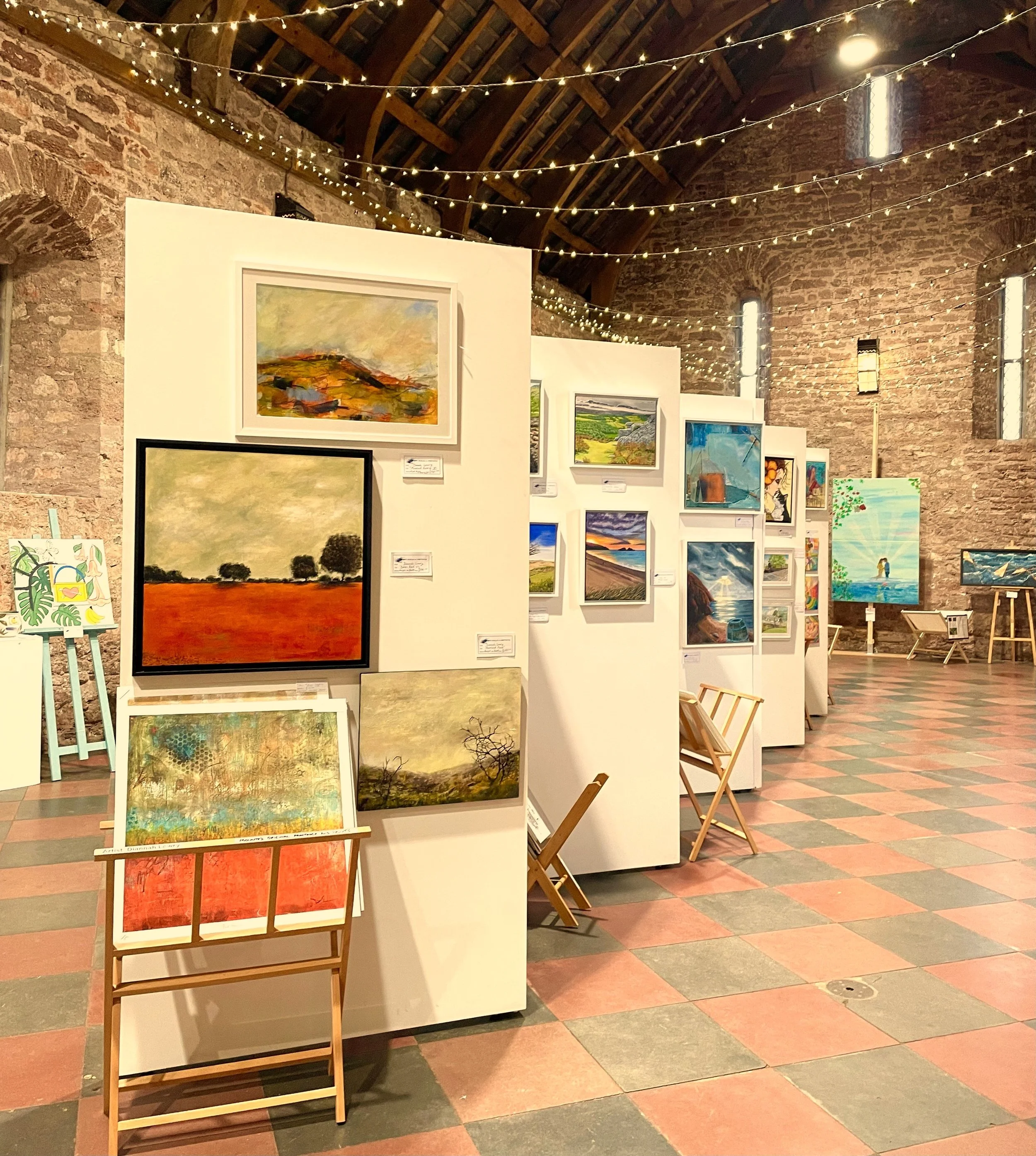 Art gallery with paintings displayed on white partitions and easels, exposed brick walls, string lights hanging from the ceiling, and colorful tiled floor.