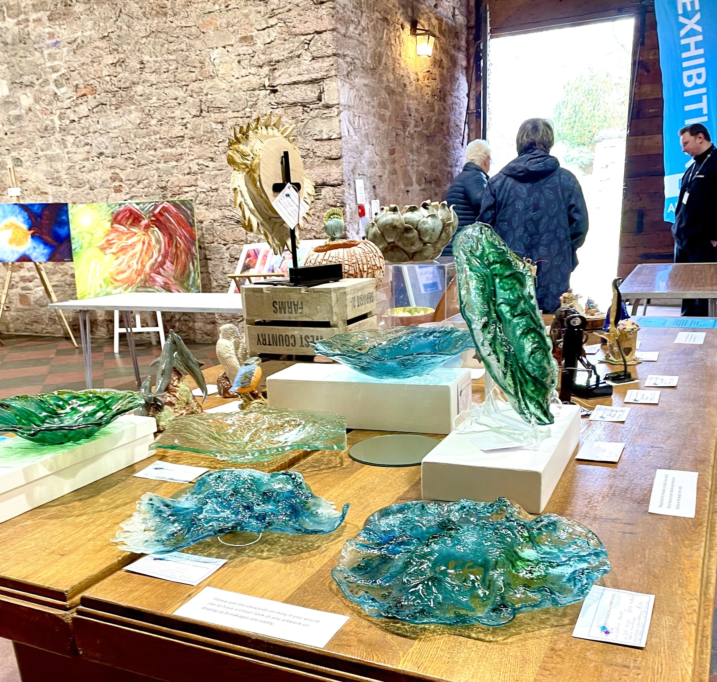 Indoor art exhibit featuring colorful glass sculptures and paintings on display against a rustic brick wall. Three people are viewing the art, with a 'Exhibit' banner on the right side of the image.