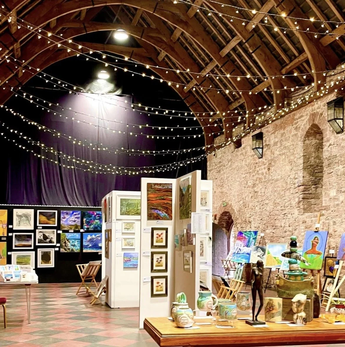 Art gallery or exhibition space with painting displays, sculptures, and decorative pottery, illuminated by string lights hanging from wooden beams inside a venue with brick walls.