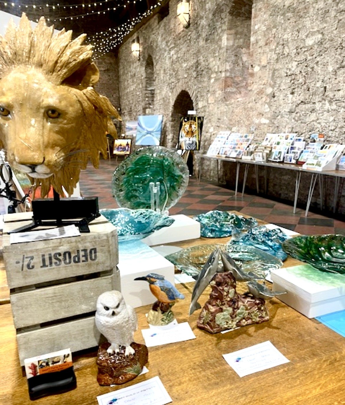 Display of glass art sculptures, animal figurines, and paintings set up in a brick-walled gallery with tables and artwork in the background.