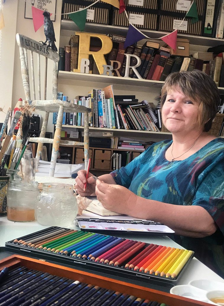 A woman sitting at a desk with art supplies, including colored pencils, paintbrushes, and jars, in a library or studio. She is smiling at the camera, wearing a colorful shirt, with books and decorative letters in the background.