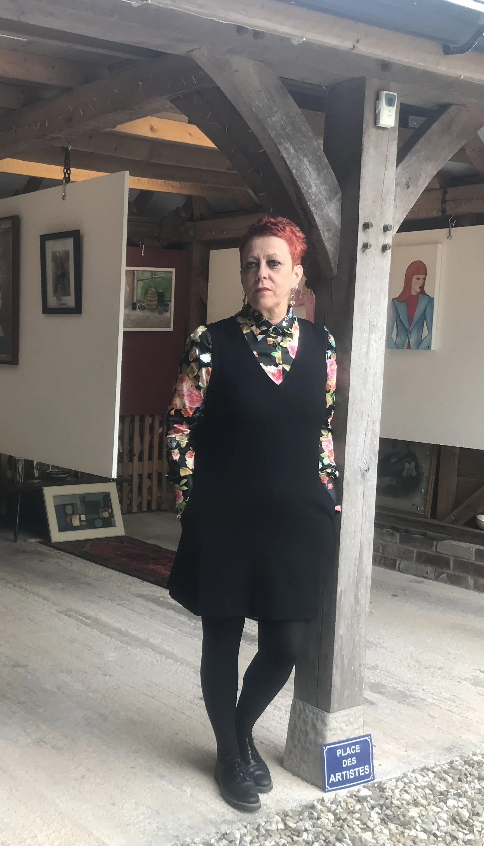 Woman with short red hair wearing a floral shirt and black dress, standing next to a wooden support beam in an art gallery, with artwork on the walls and a small sign reading "Place des Artistes".