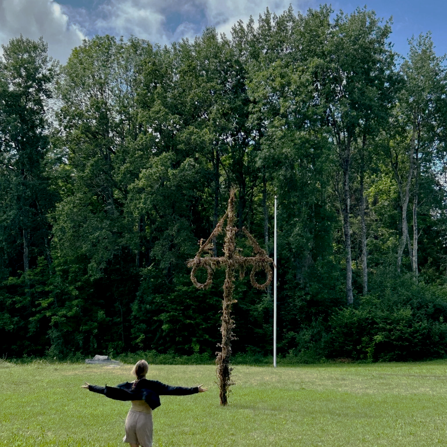 A person standing on grass with arms outstretched towards a large decorative cross made of natural materials, against a backdrop of trees and a partly cloudy sky.